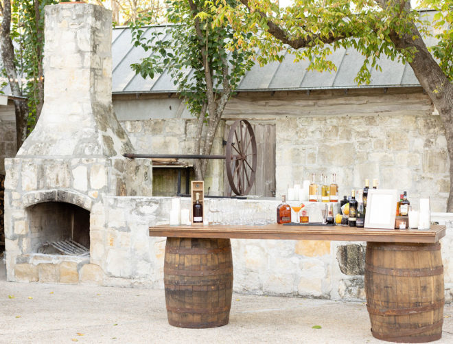 Outdoor fireplace and wine barrel table set with alcohol bottles, cocktail glasses and a drink menu in a 1800s style courtyard event space at Kendall Hill Country Inn in Boerne. 