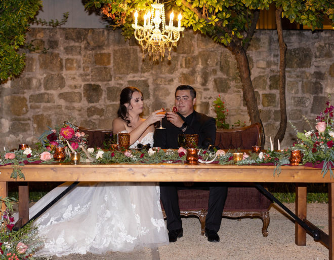 Bride toasts a cocktail glass against a suited grooms tumbler while seated at a long farmhouse table set with candle holders, greenery and pink and red flowers underneath a hanging chandelier in the courtyard of The Kendall Hill Country Inn in downtown Boerne. 