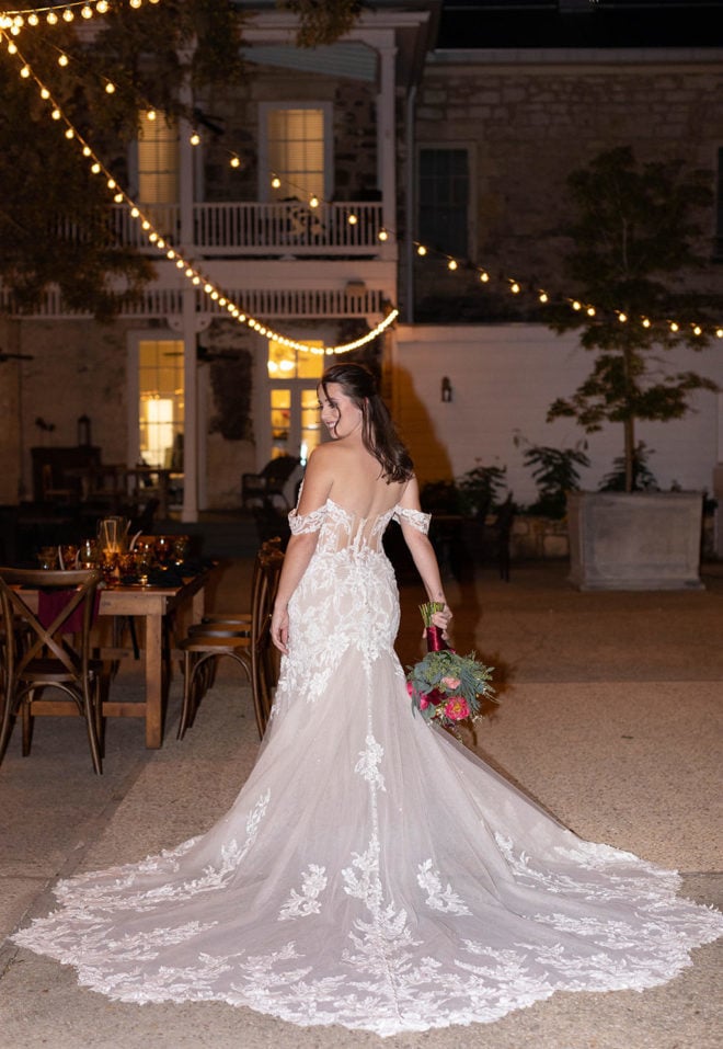 Women in long floral applique wedding gown holding bouquet of florals stands in the center of the courtyard with lit string lights strung above her at The Kendall Hill Country Inn. 