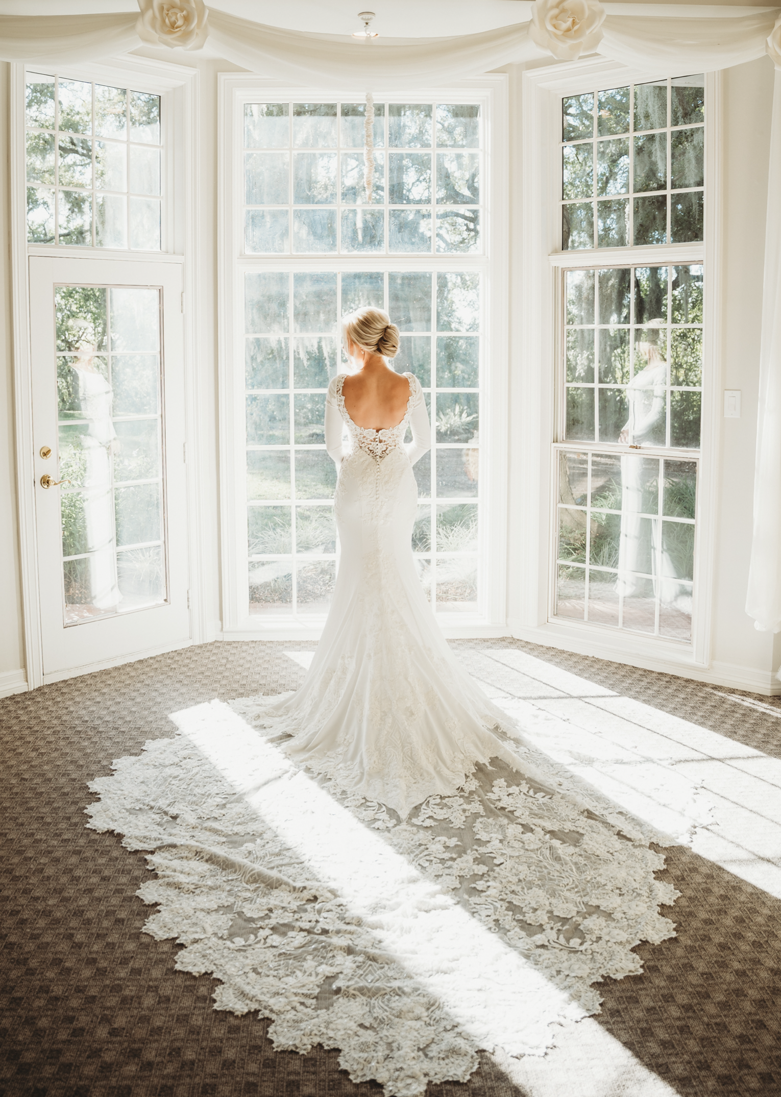 The bride stands by the window why sunlight hits her wedding gown.