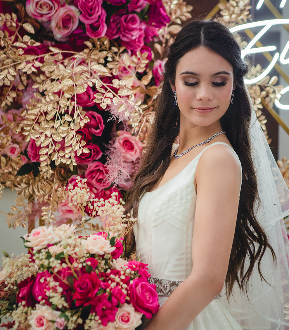 A bride is wearing gold sparkly eye shadow and her long brown hair is losely curled and half pulled back. She is holding a pink and gold bridal bouquet with flowers behind her. 