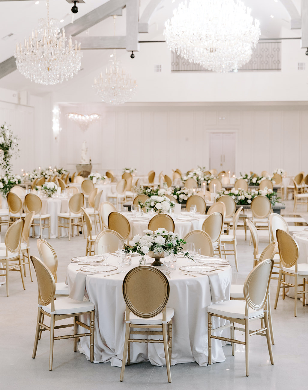 The reception room is decorated and ready for the wedding party and guests. Crystal chandeliers hang from the ceiling and lush greenery and ivory blooms fill the centerpieces on the tables.