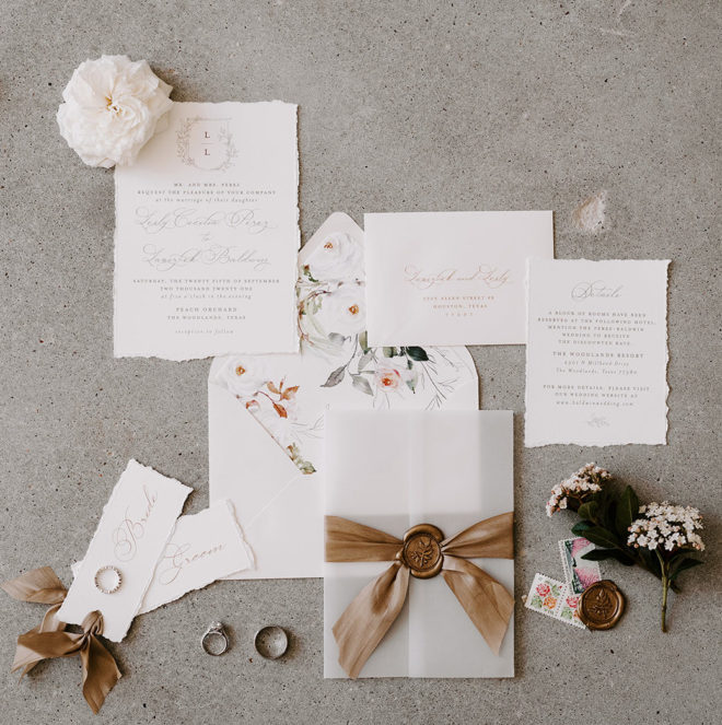 Flatlay with an invitation suite featuring bronze ribbons, rings, ivory blooms and cursive script at a wedding at the Peach Orchard Venue. 