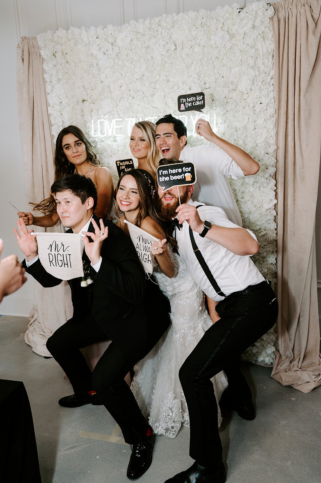 The bride and groom pose with their guests in front of a photo backdrop full of white flowers and a neon sign at their wedding reception.