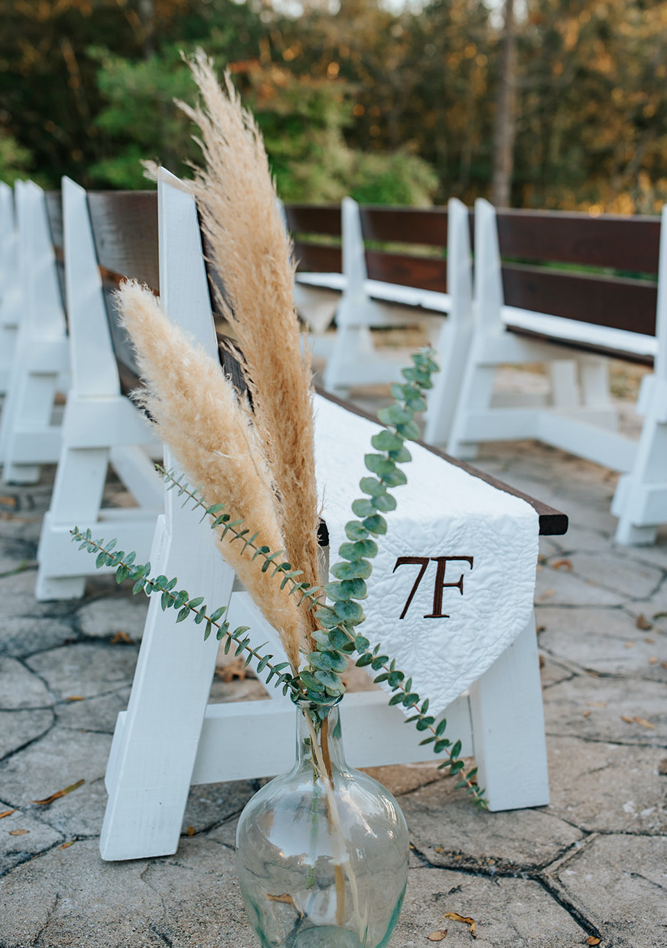 A glass bottle filled with Pampas grass and Eucalyptus is placed next to a white aisle bench outside for a winter wedding ceremony at 7F Lodge in College Station, TX.