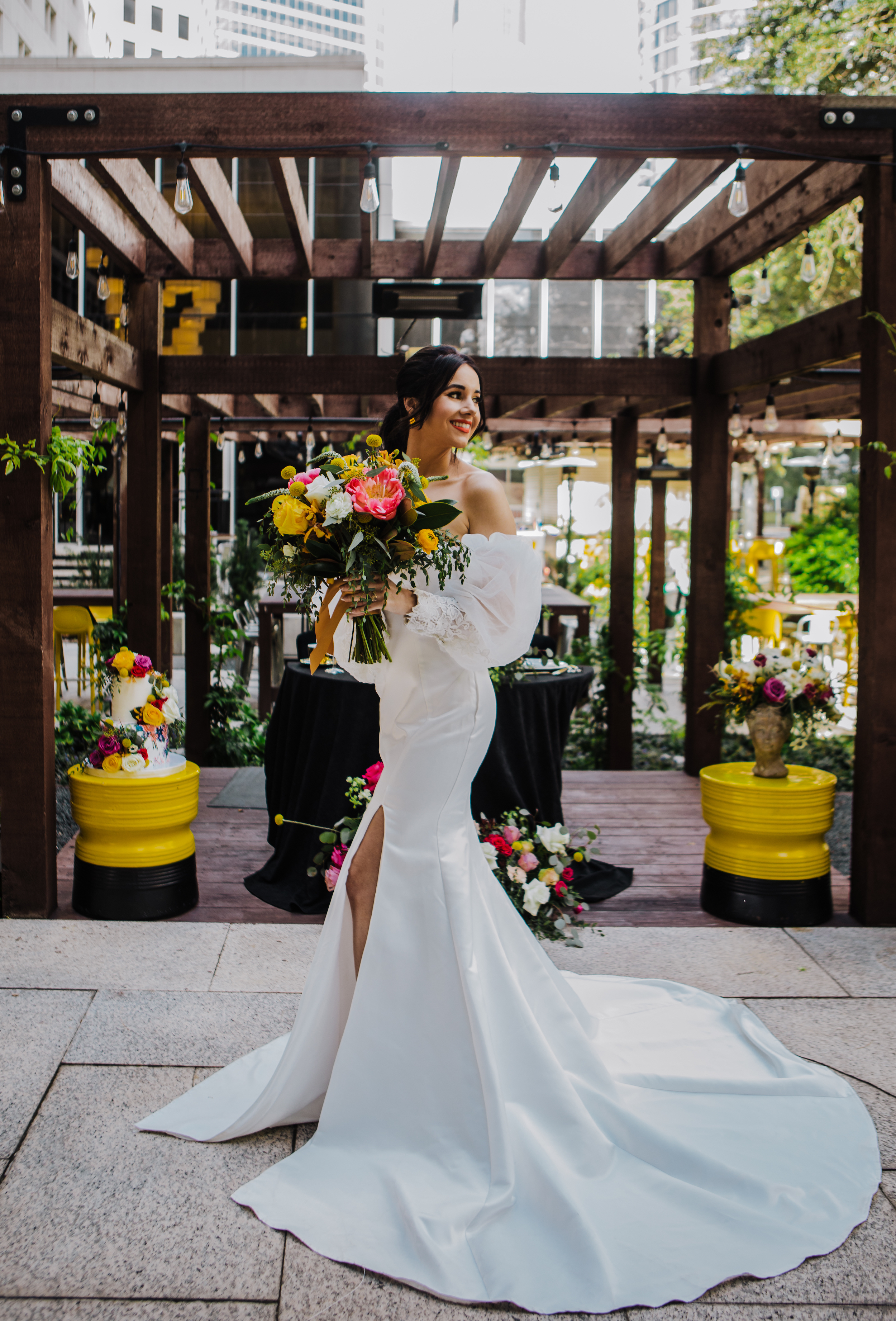 A bride smiles in a sleek off-the-shoulder gown by Kary King Boutique, holding a colorful bridal bouquet with yellow and pink flowers on the outdoor courtyard at a downtown Houston wedding venue.