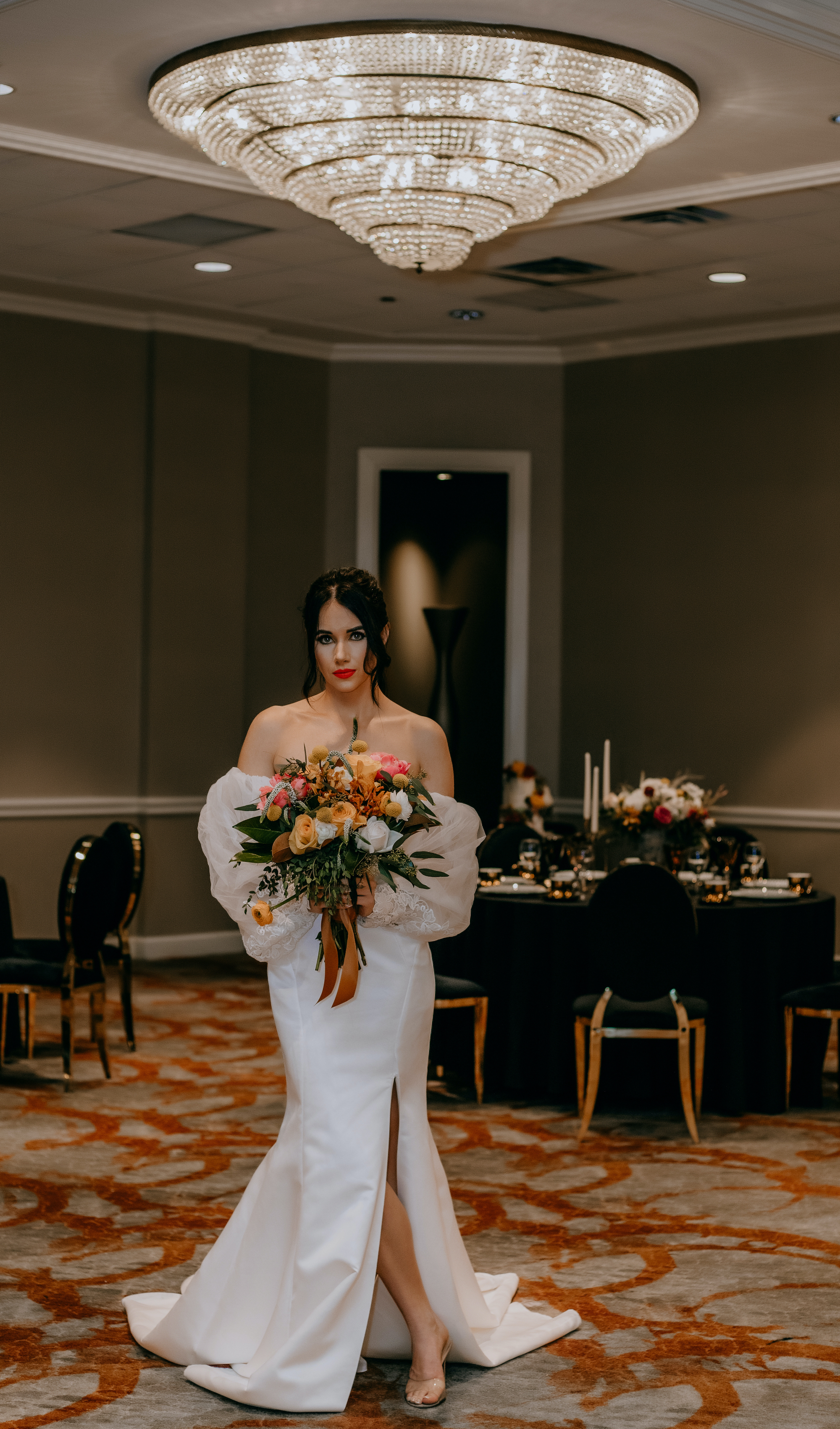 A bride stands under a crystal chandelier holding a vibrant bridal bouquet at The Whitehall in Houston, TX. 