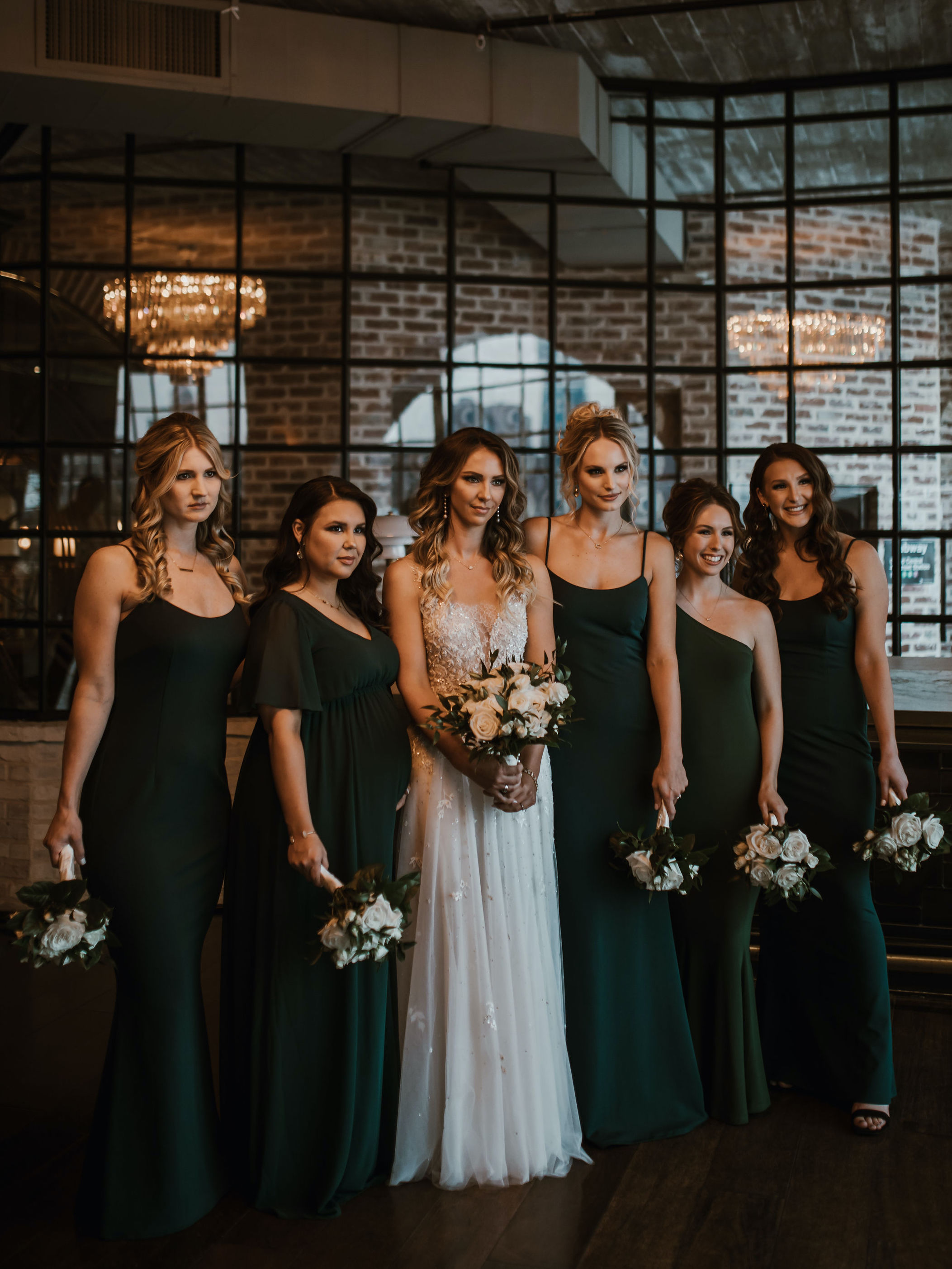 The bride poses with her bridesmaids who are all wearing emerald green dresses and white flower bouquets with greenery. They are standing in The Astorian, a Houston wedding venue.
