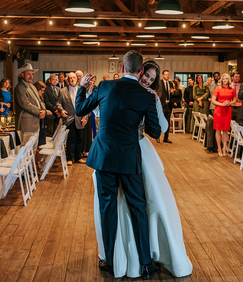 The bride and groom dance as their wedding guests watch from their seats. They are in a rustic reception room at 7F Lodge in College Station, TX.
