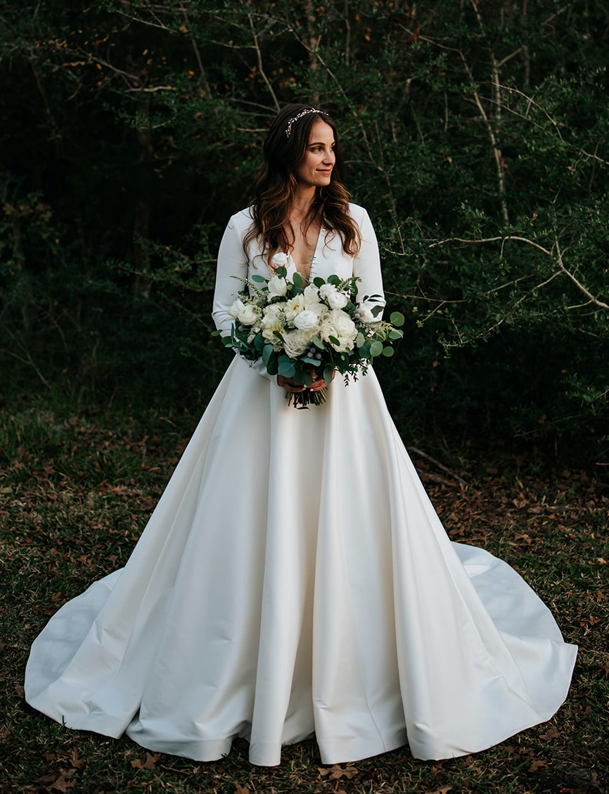 The bride stands near the woods holding her ivory bridal bouquet and wearing a white long sleeve bridal gown for her winter wedding at 7F Lodge in College Station, TX.