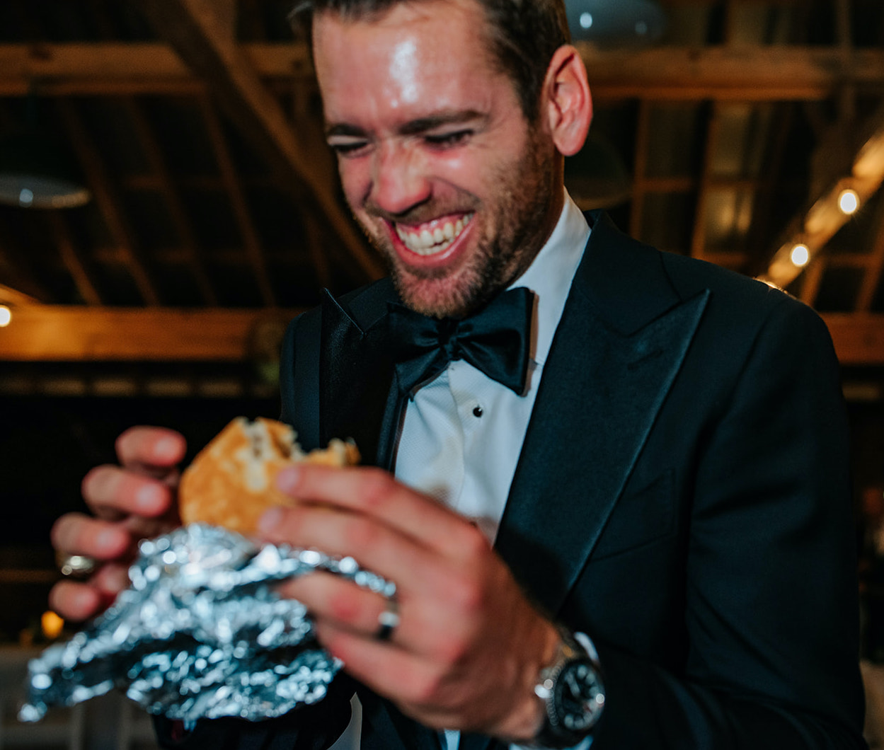 The groom laughs while holding a chicken biscuit from Whataburger at their winter wedding reception at 7F Lodge in College Station, TX.