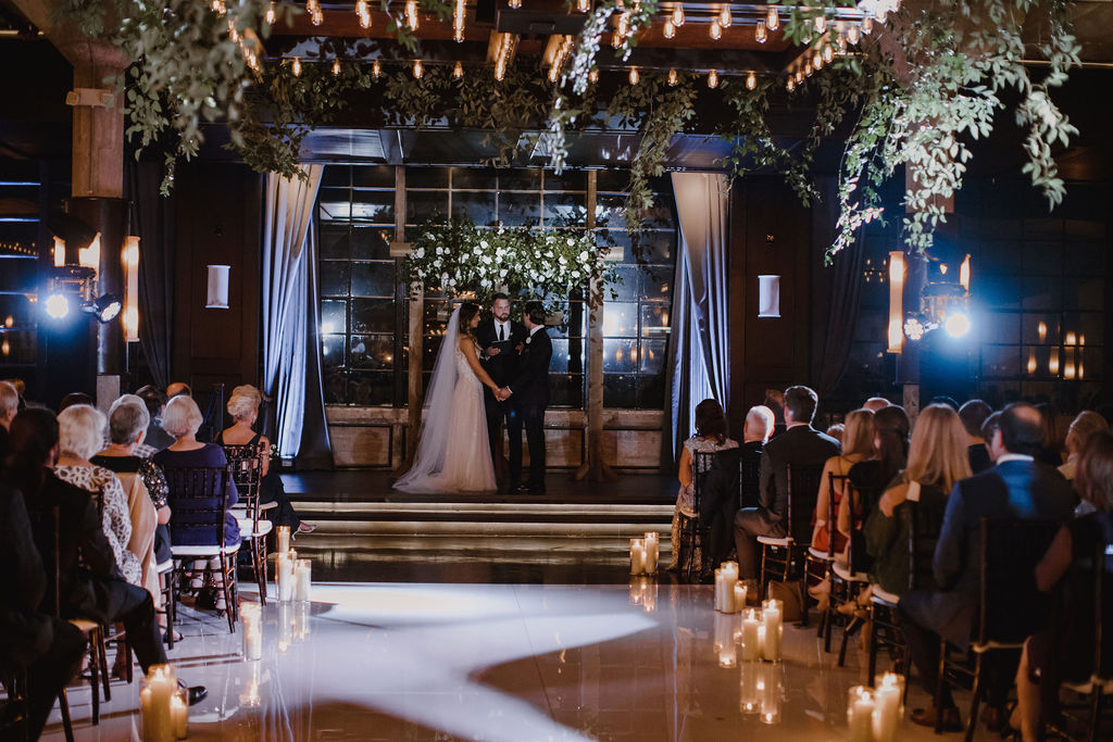 The bride and groom stand at the altar holding hands while their guests watch beneath a canopy of greenery and a candlelit aisle. The couple celebrates their love story at The Astorian, a wedding venue in Houston, TX.