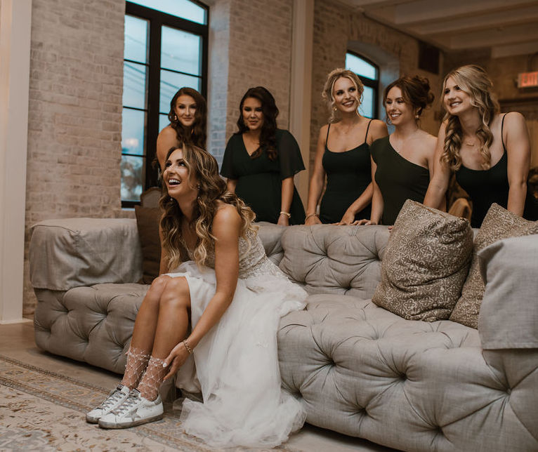 The bride puts on her white Golden Goose shoes and smiles while her bridesmaids stand behind her. They are in the bridal suite at The Astorian, a wedding venue in Houston,TX.