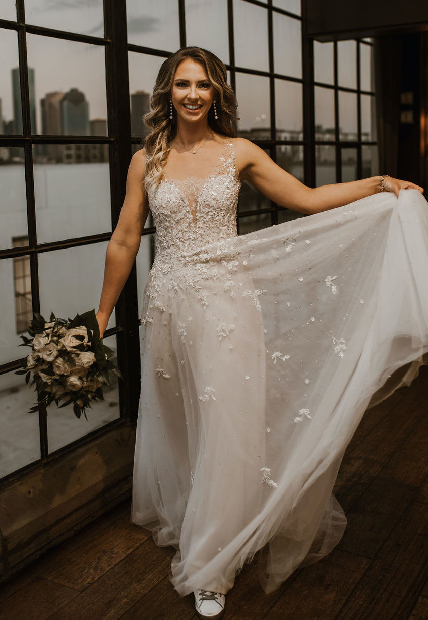 The bride smiles and lifts her wedding dress skirt while she walks forward holding her bouquet. The Houston skyline can be seen from the window close to her as she is about to celebrate her love story at The Astorian.