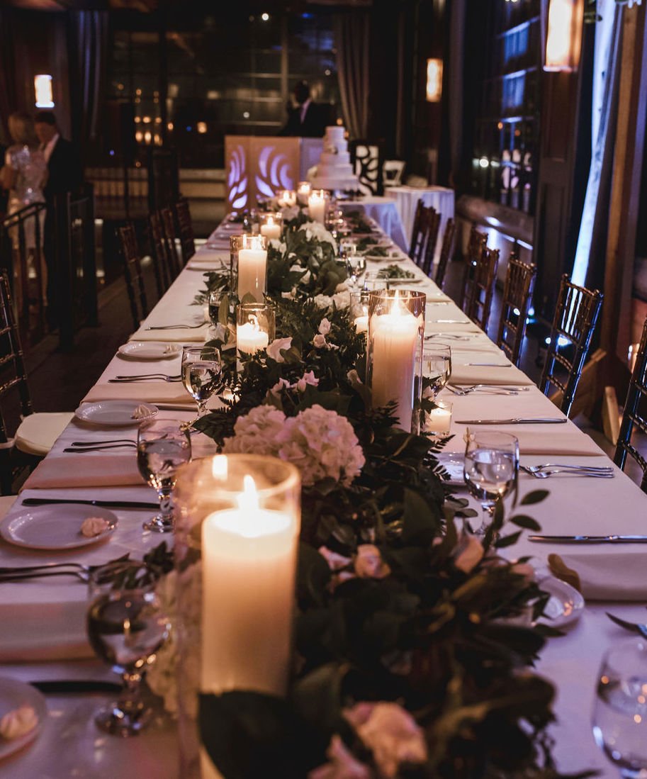 A long table is set for a wedding reception with candles, white flowers and greenery at The Astorian in Houston, Texas.