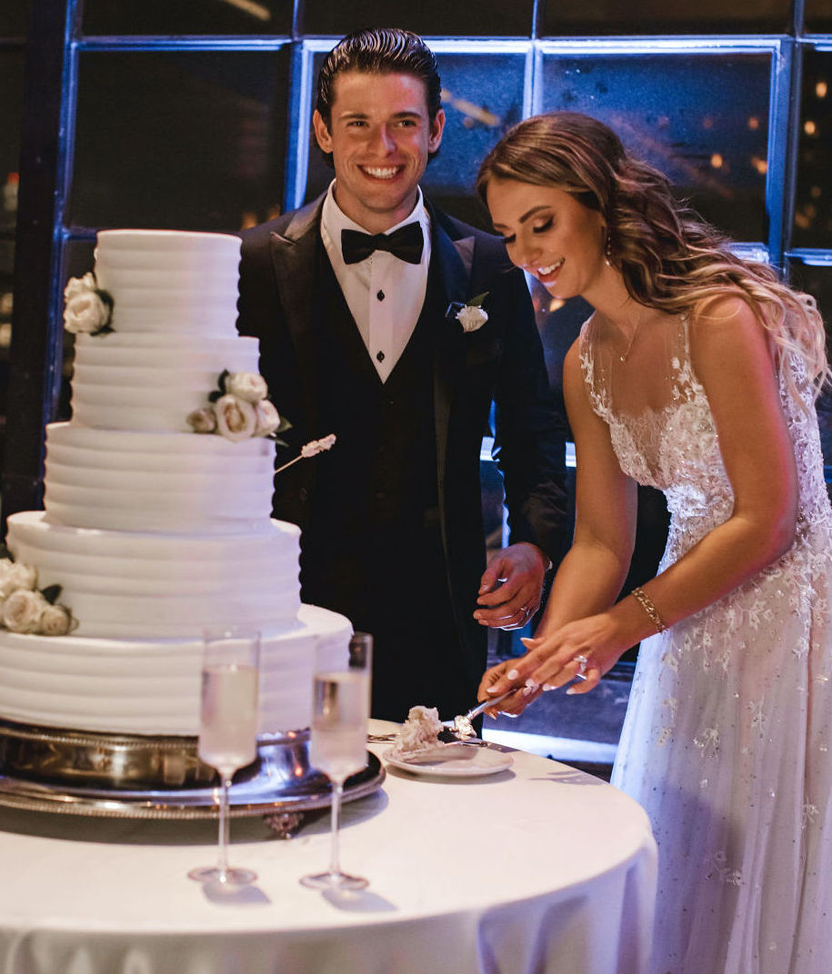 The bride and groom stand next to their five-tier white wedding cake as the bride cuts a piece. Their reception is at The Astorian in Houston, TX.