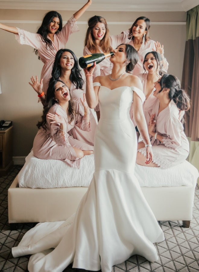 Bride drinks a bottle of champagne in her wedding gown while bridesmaids in robes smile and laugh behind her in a hotel room at the San Luis Resort, Spa and Conference Center. 