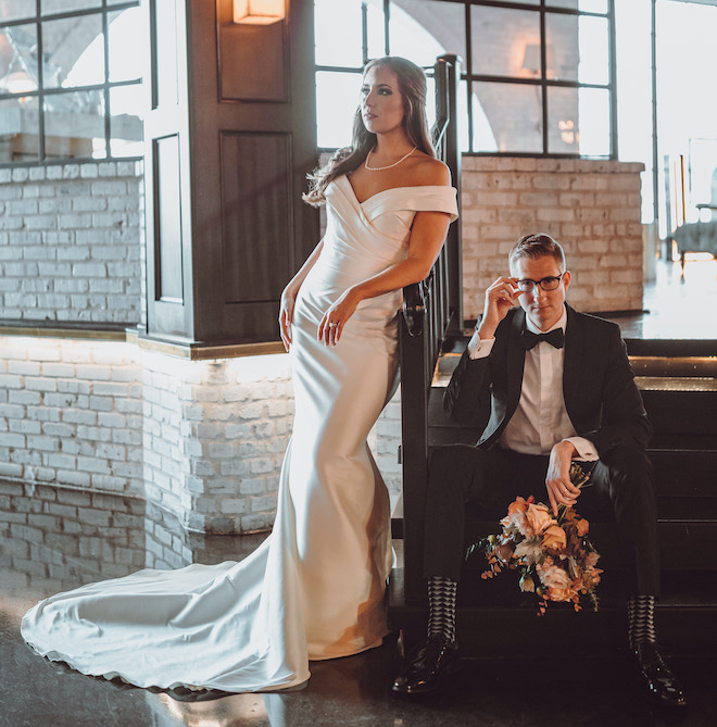 Bride leans on railing while her husband in a black suit and glasses sits on the stairs during their wedding reception at Houston wedding venue, The Astorian. 