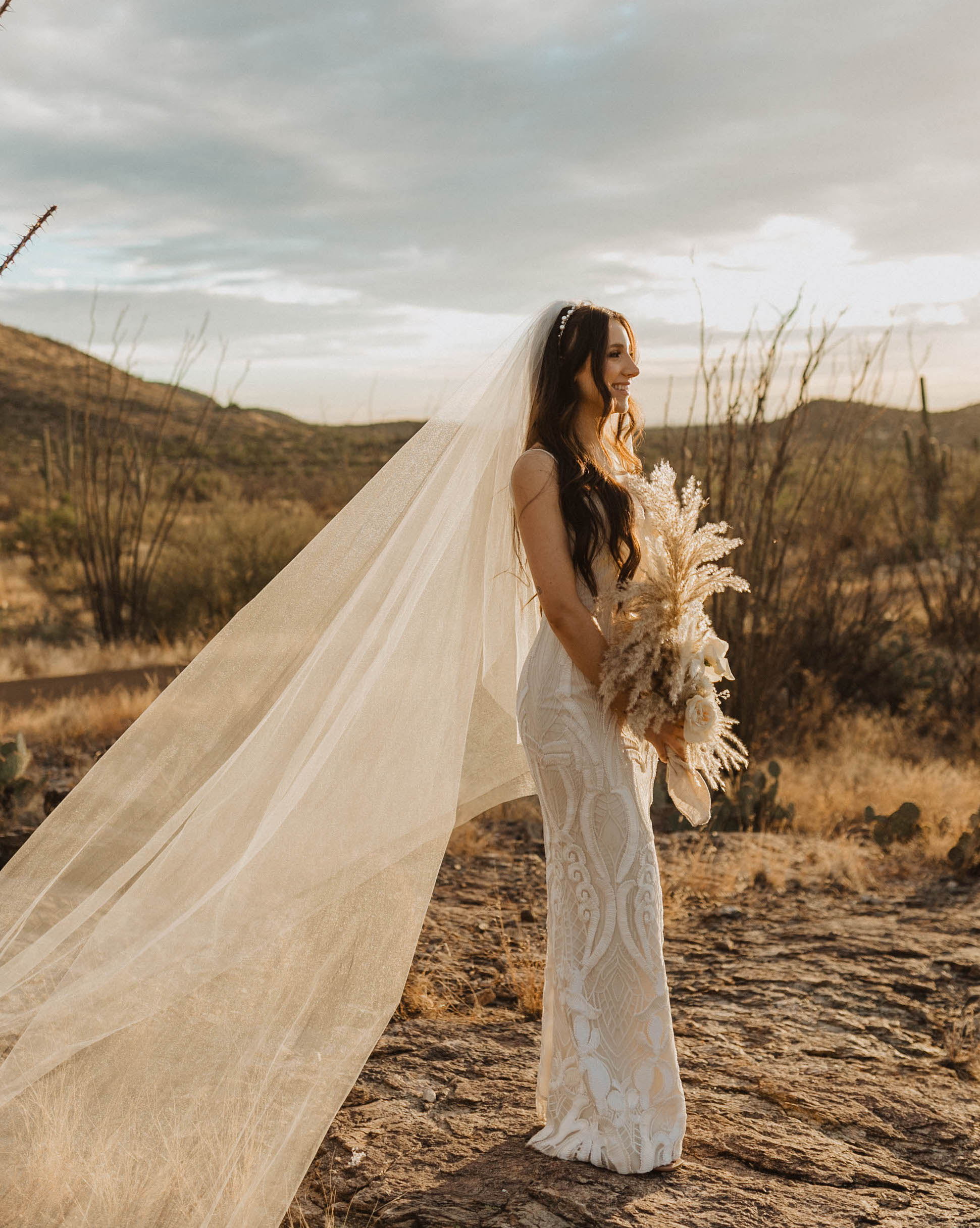 The bride (Caked Up With Riley) smiles outside in the desert sun at Saguaro National Park in Arizona. She holds a neutral bohemian style bridal bouquet.