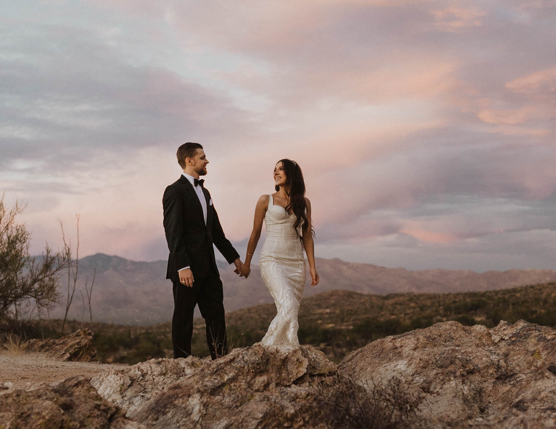 The bride and groom hold hands outside in Arizona with a sunset behind them.
