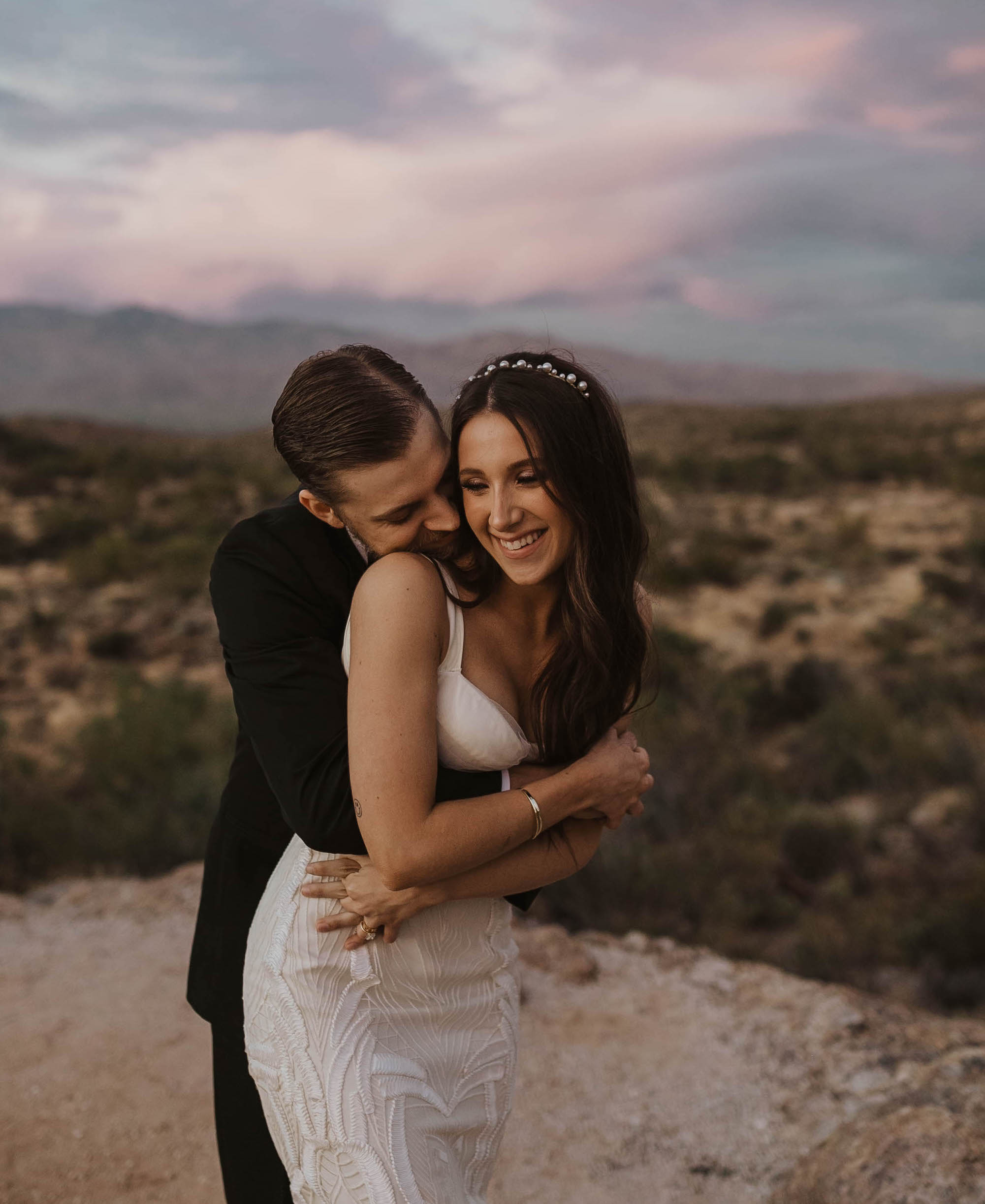 A groom hugs his bride from behind as they smile. The sunset is beautiful at their elopement in Arizona.