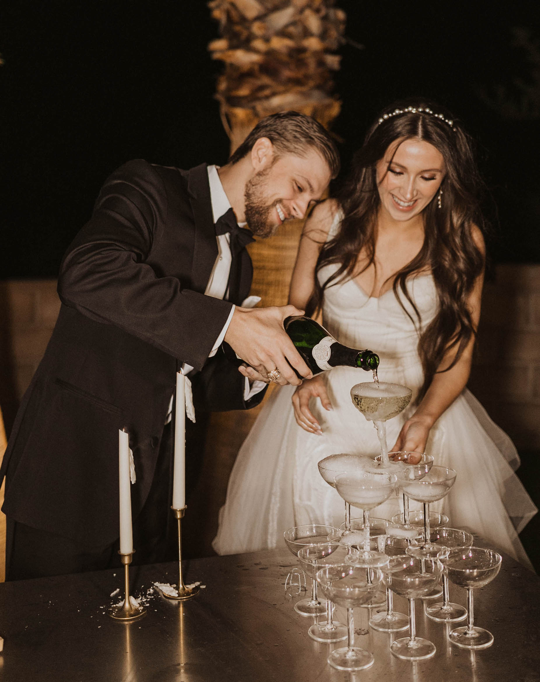A bride and groom smile and pour champagne into a coupe tower at their wedding reception.