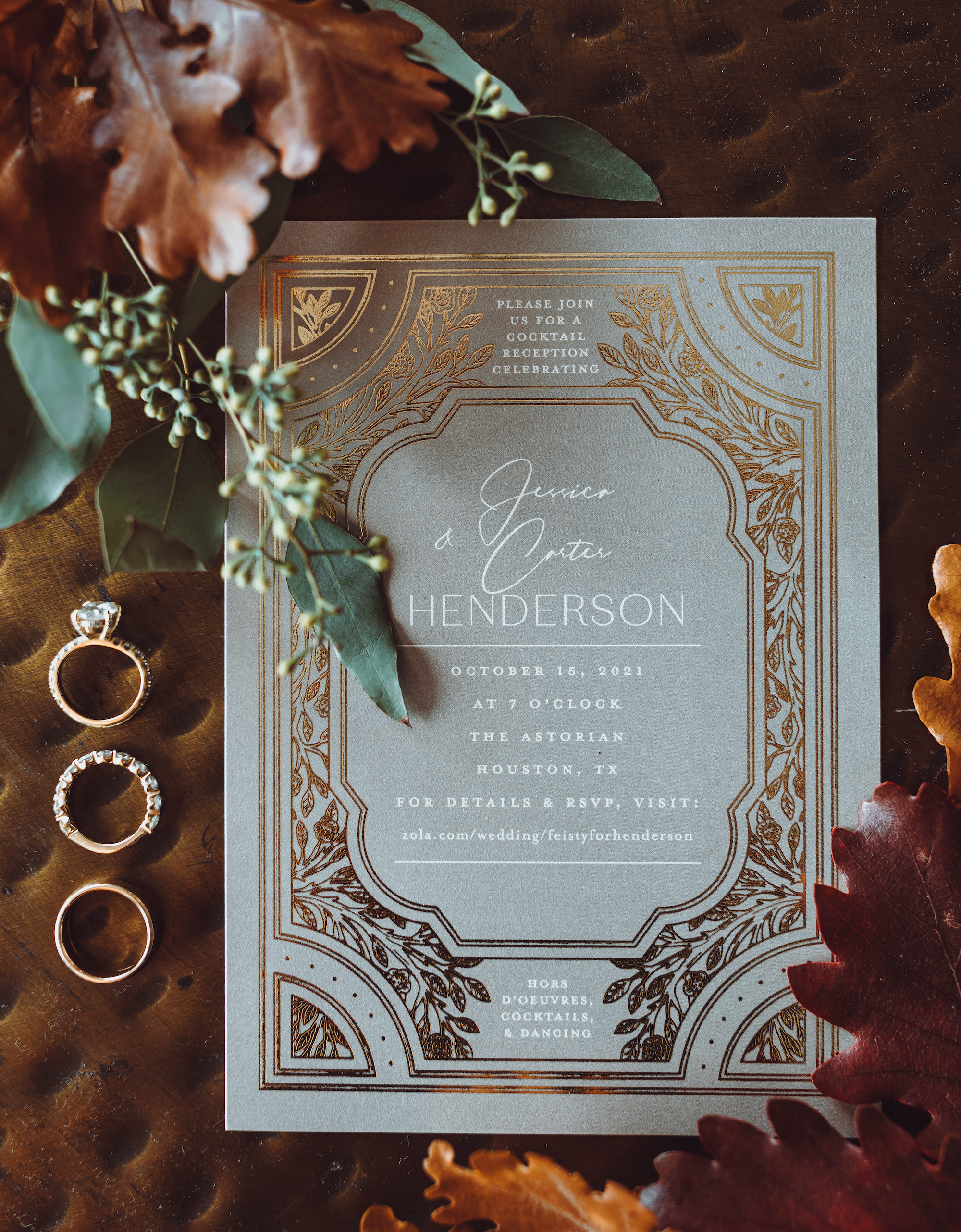 A gold-engraved wedding invitation with fall leaves surrounding it and engagement rings on one side.