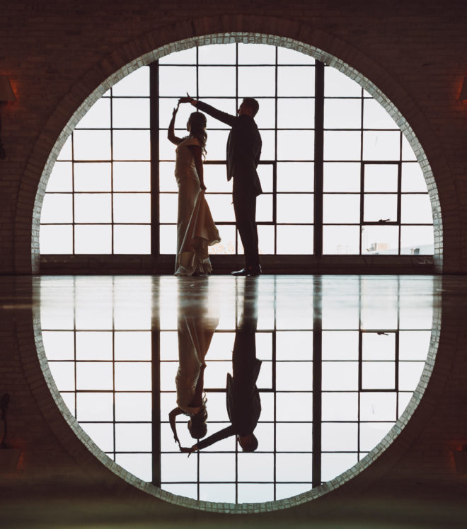 Bride and groom dance in front of a large half circle window inside industrial chic wedding venue, The Astorian, in Houston, Texas.