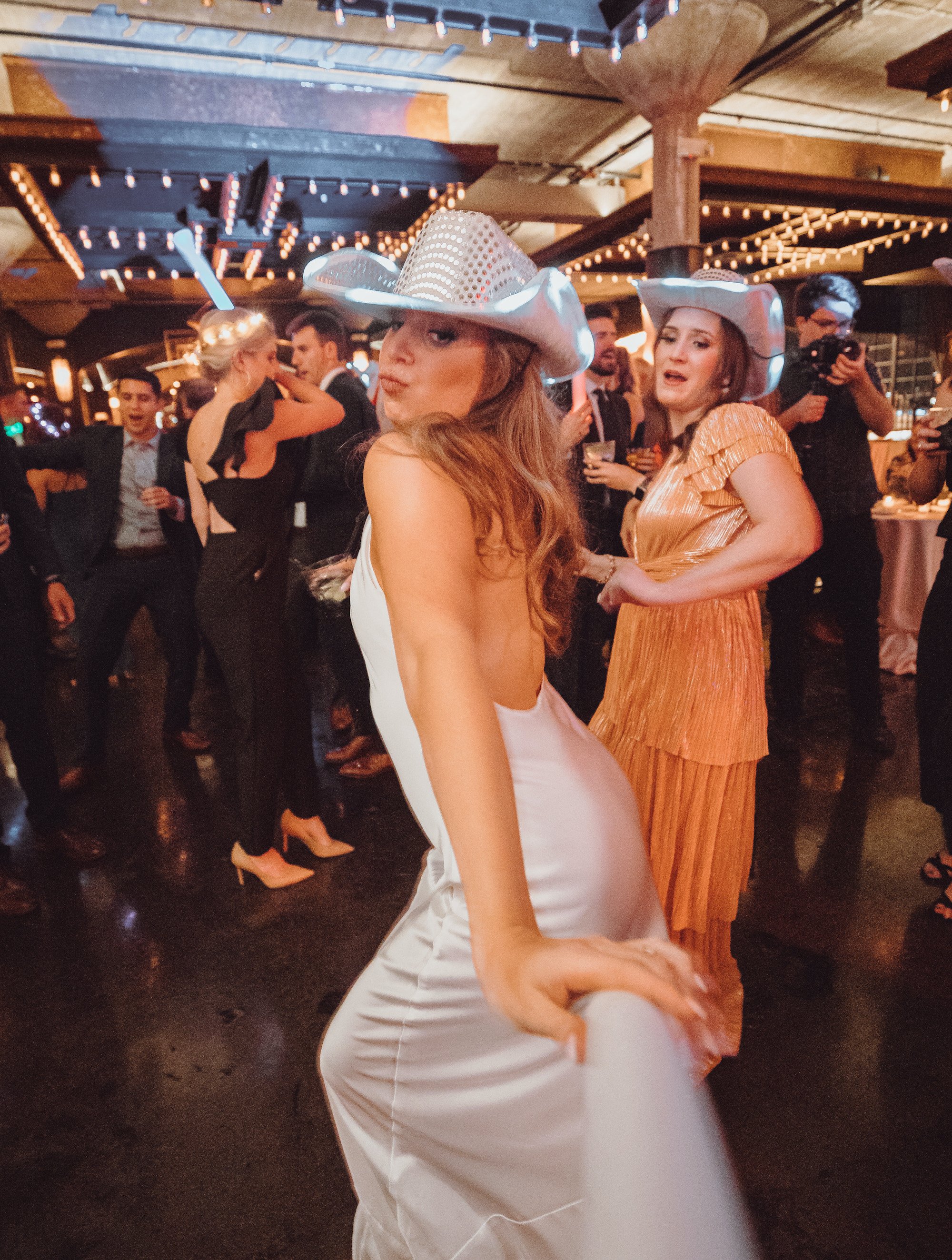 Bride in white silk slip dress and cowboy hat dances at her wedding reception inside Houston wedding venue, The Astorian. 
