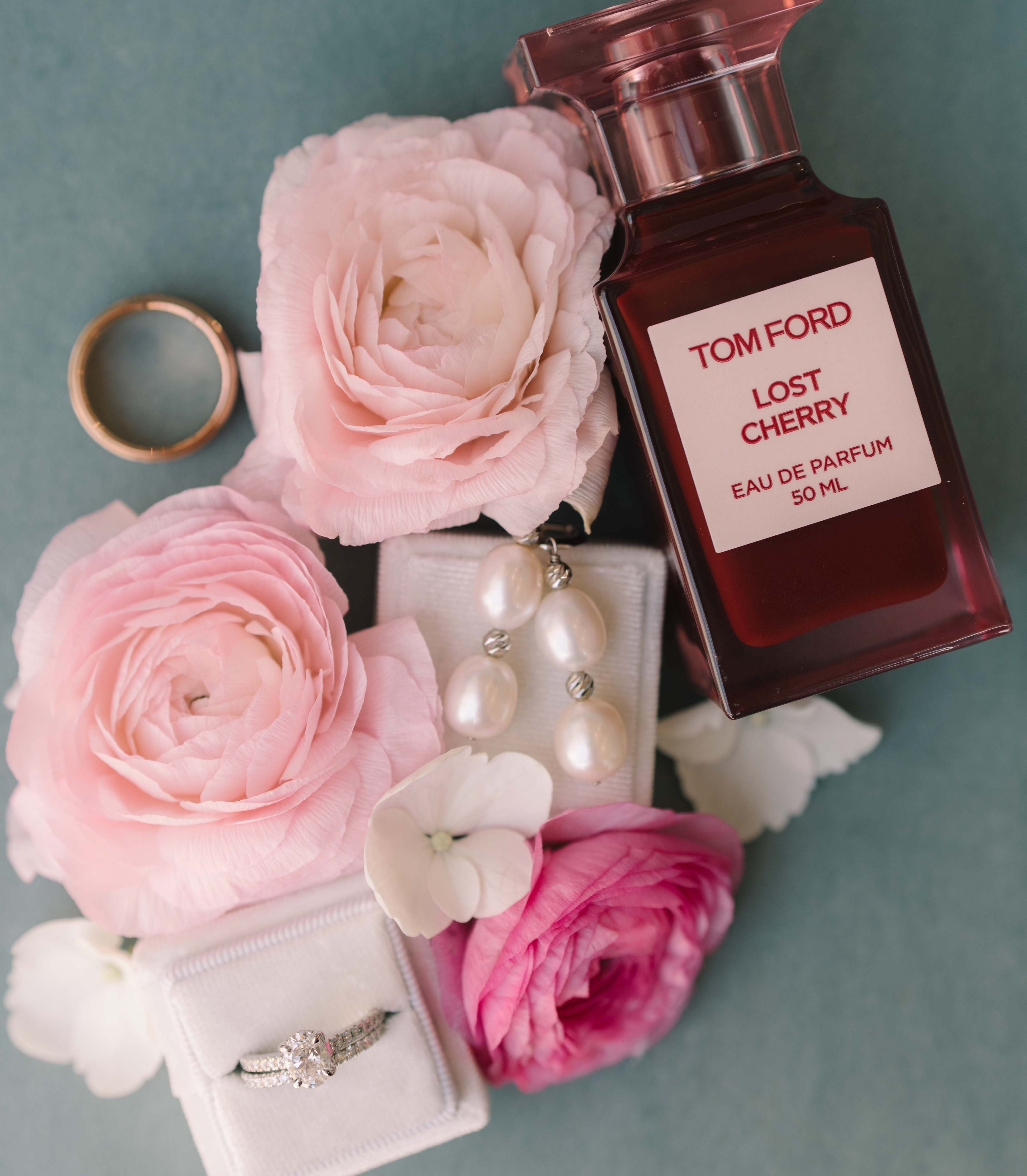 A flatlay full of pink flowers, Tom Ford "Lost Cherry" perfume and pearl earrings with an engagement ring.