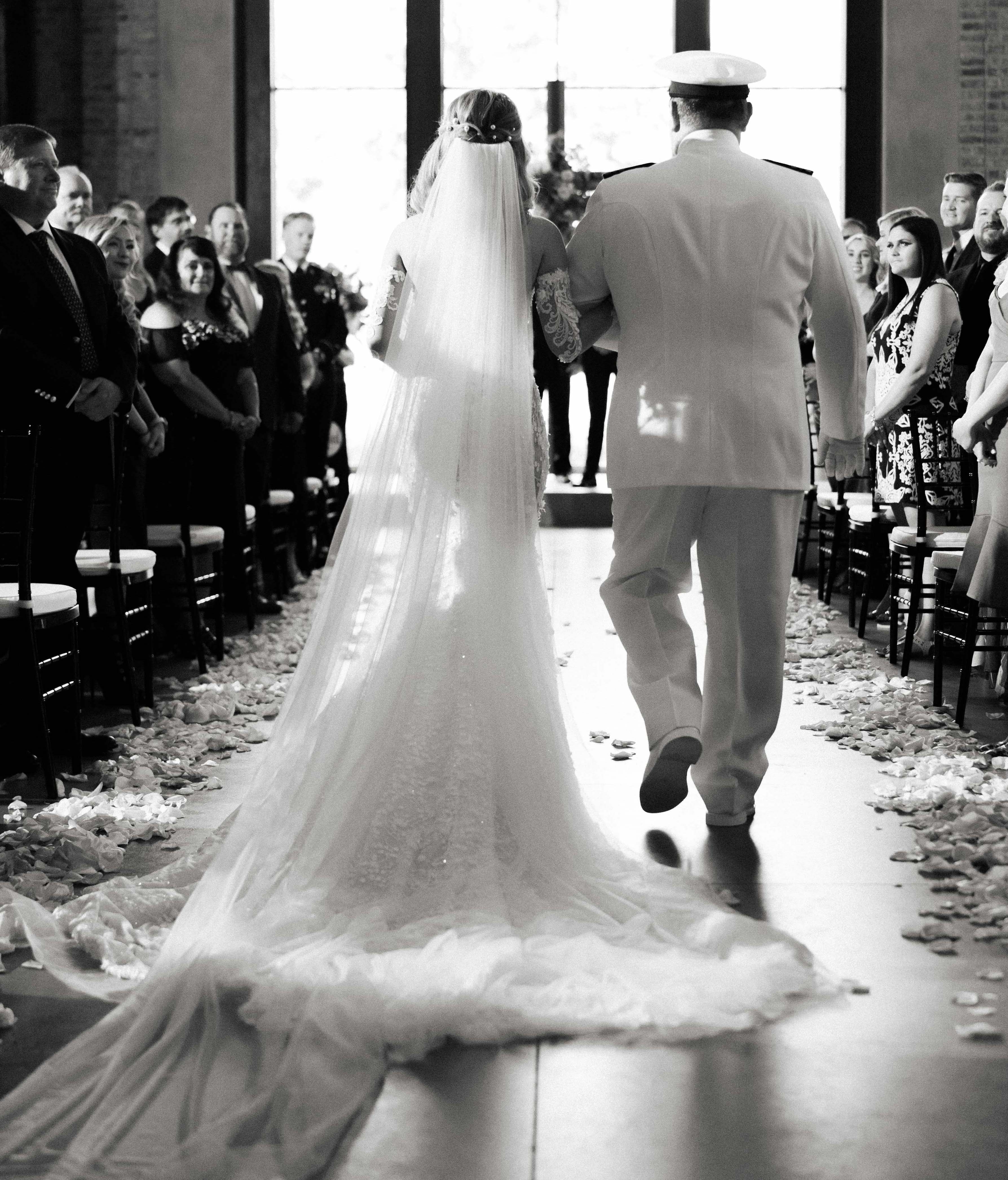 A black and white photo of a bride walking down the aisle with her dad. Their backs are turned to the camera.