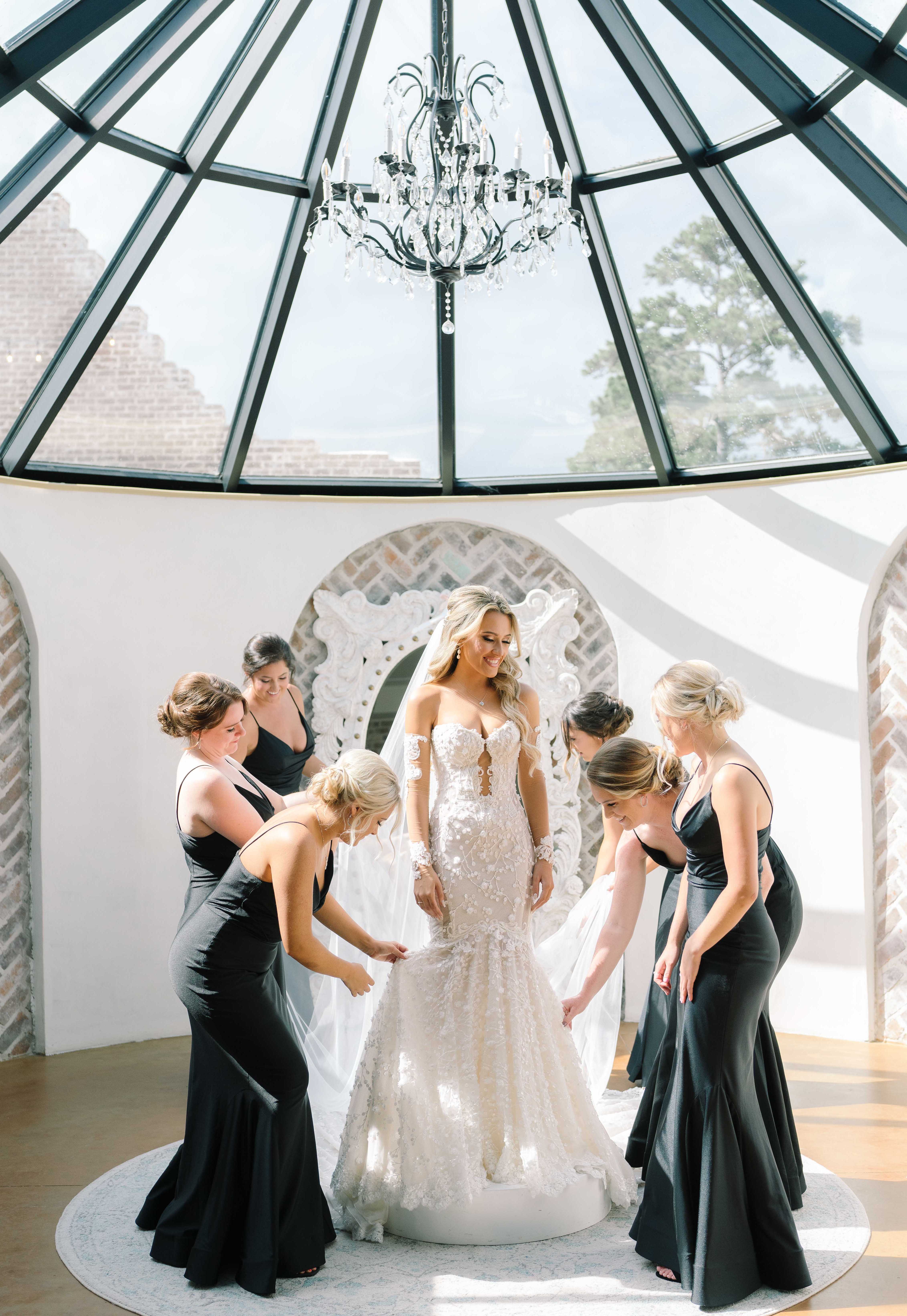 A bride stands in a sunny bridal suite while her bridesmaids fix her wedding gown.