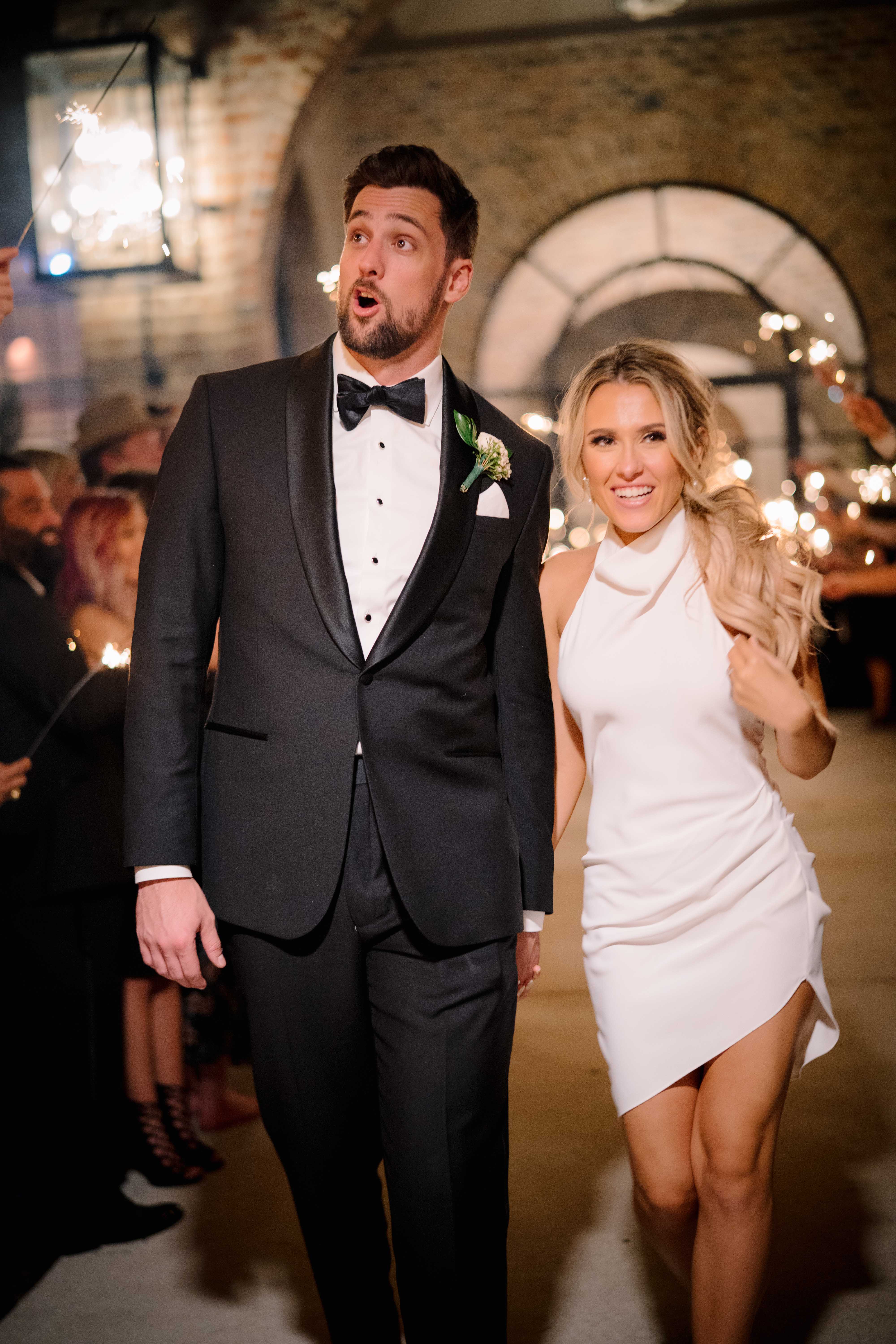 A bride and groom make their grand exit from their wedding while their wedding guests hold sparklers.