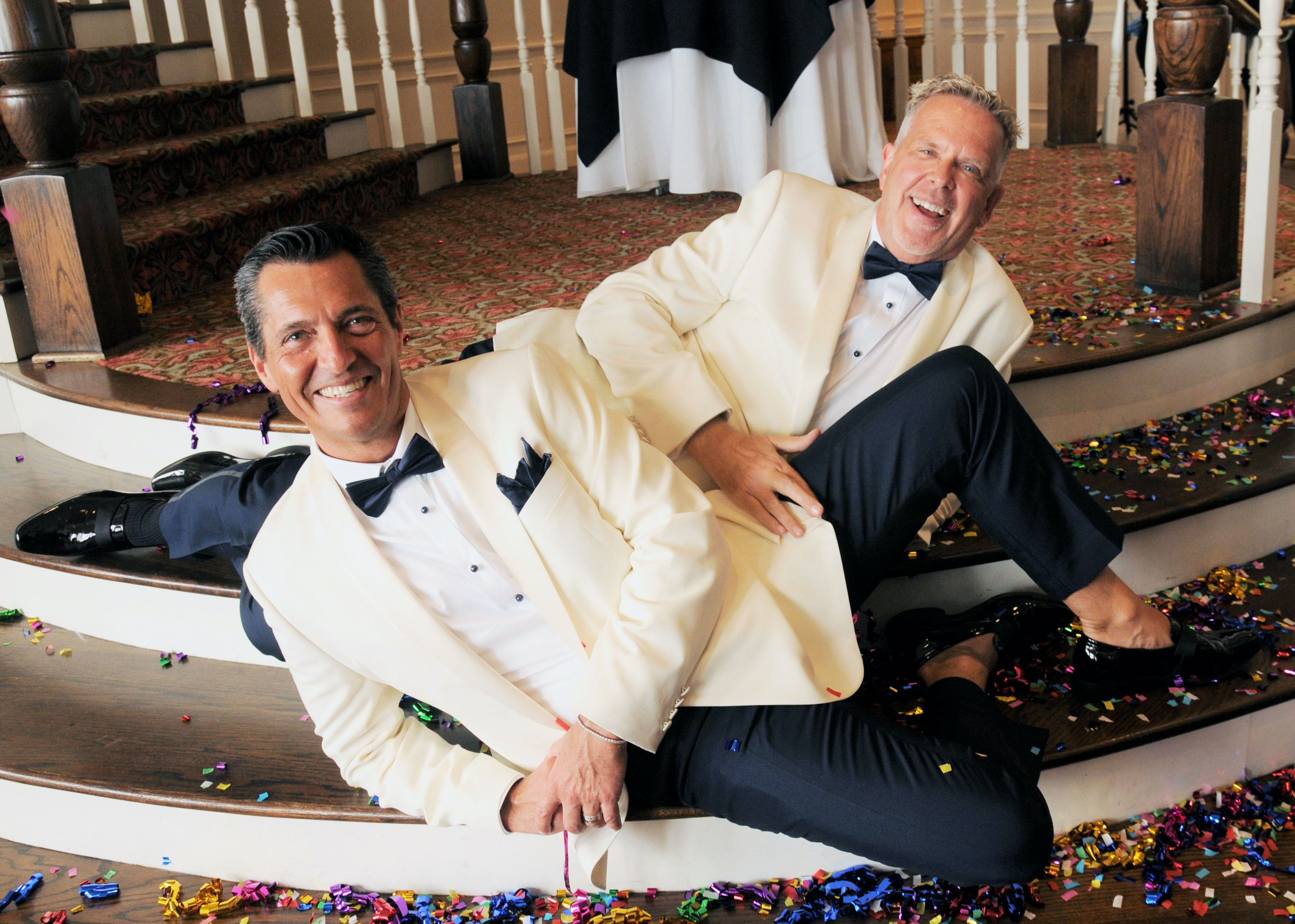 A groom and groom pose on a grand staircase with colorful confetti surrounding them on the floor.