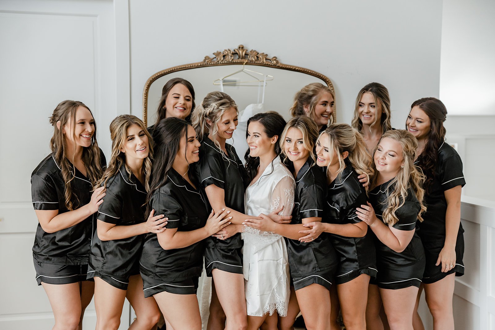 A bride laughs with her bridesmaids. They all are wearing black pajamas and the bride is in a white silky robe.