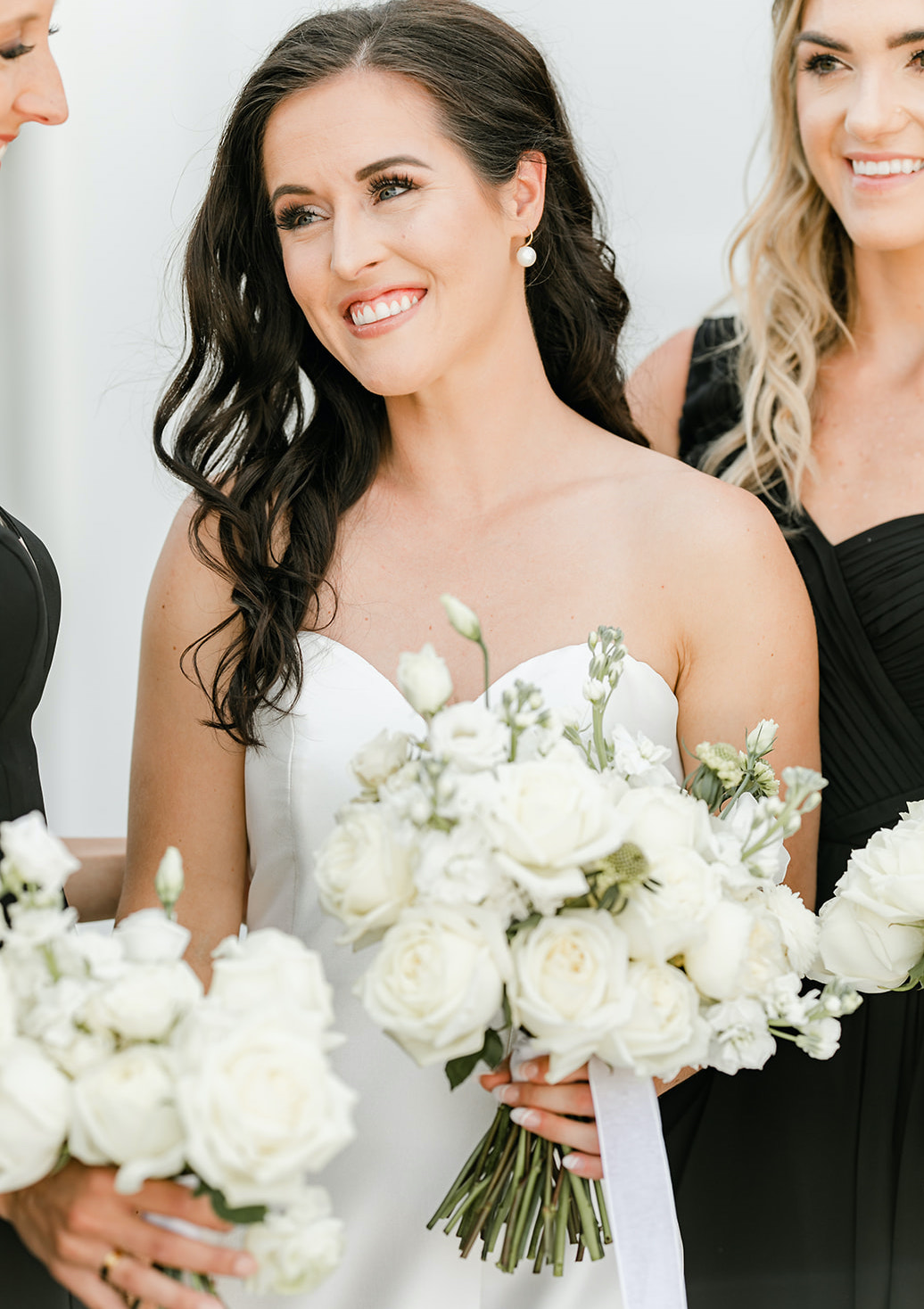 A bride smiles with her bridesmaids and holds her flower bouquet full of white roses.
