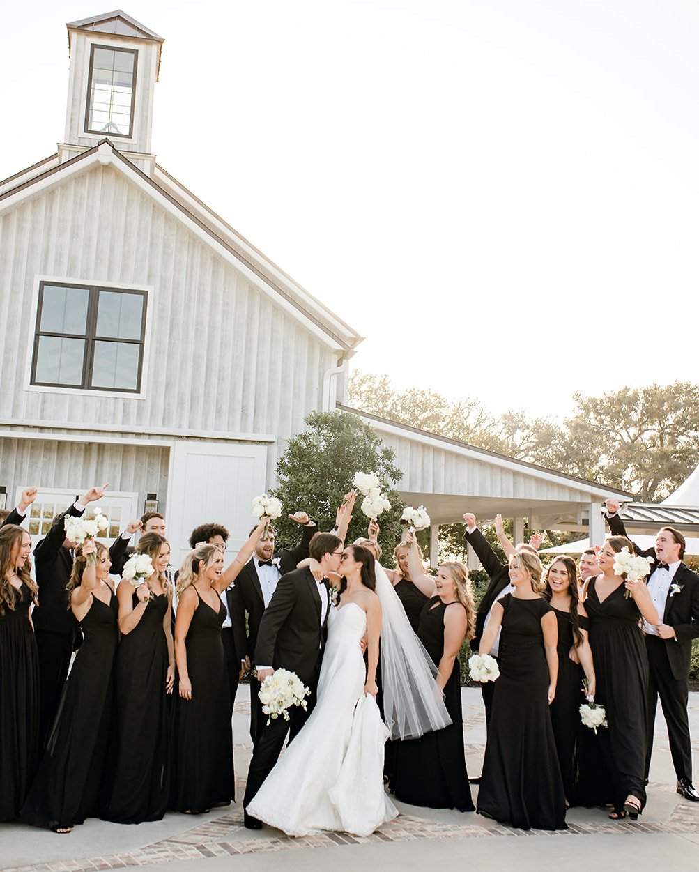A bride and groom kiss with their wedding party celebrating behind them at their elegant summer wedding in Montgomery, TX.