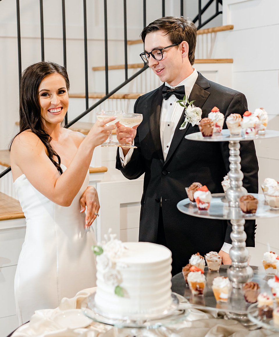 A bride and groom clink their champagne glasses before cutting into their white wedding cake.