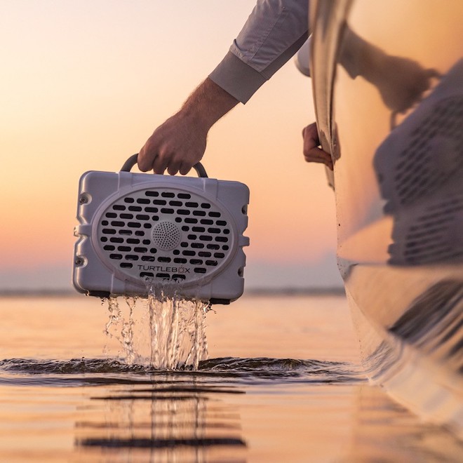 Hand holding a White Turtlebox Gen 2 Bluetooth Outdoor Speaker above a body of water. Unique grooms gifts
