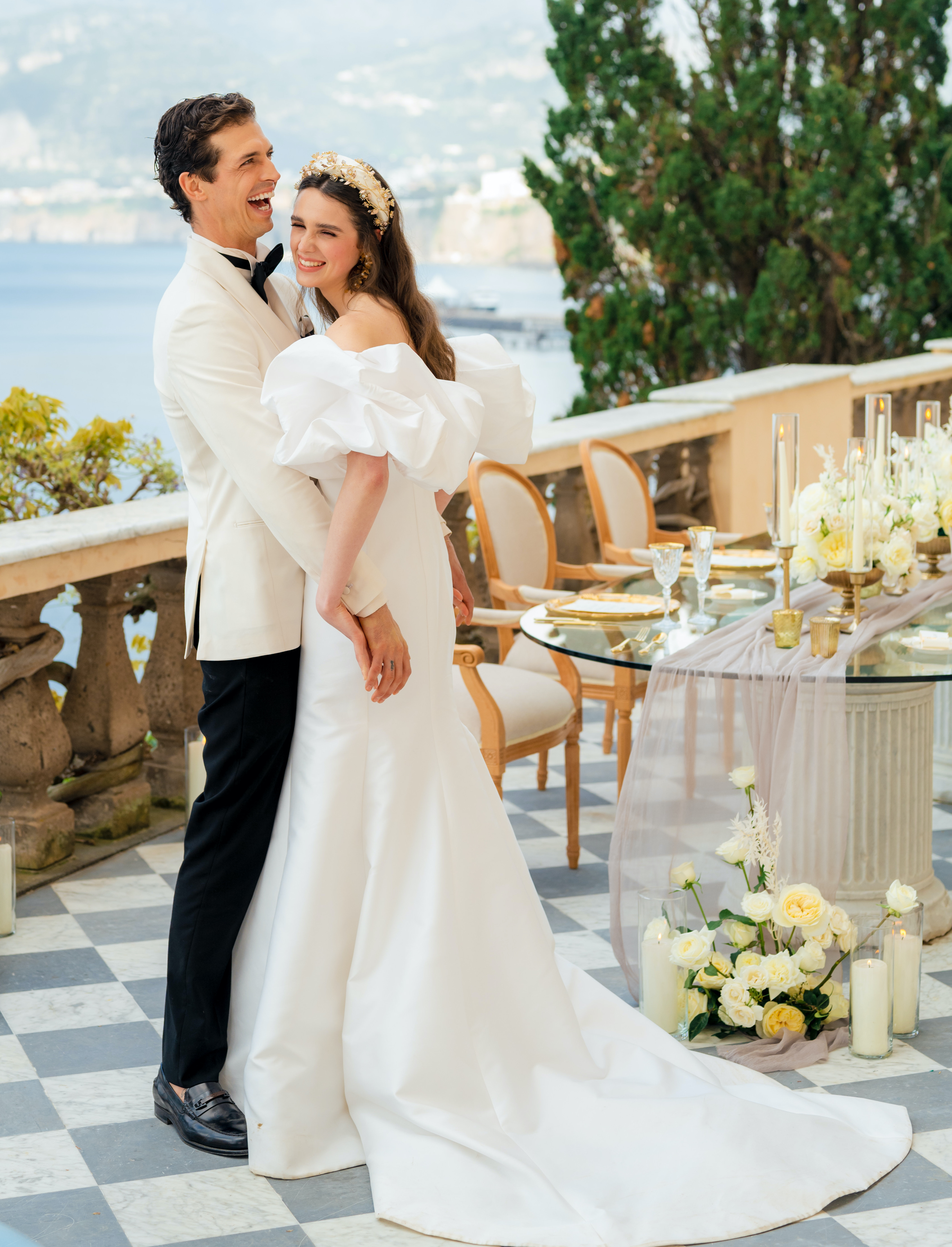 A bride and groom hold hands and laugh while standing near their alfresco reception dinner table which is full of white flowers and candles for their wedding on the Amalfi Coast.