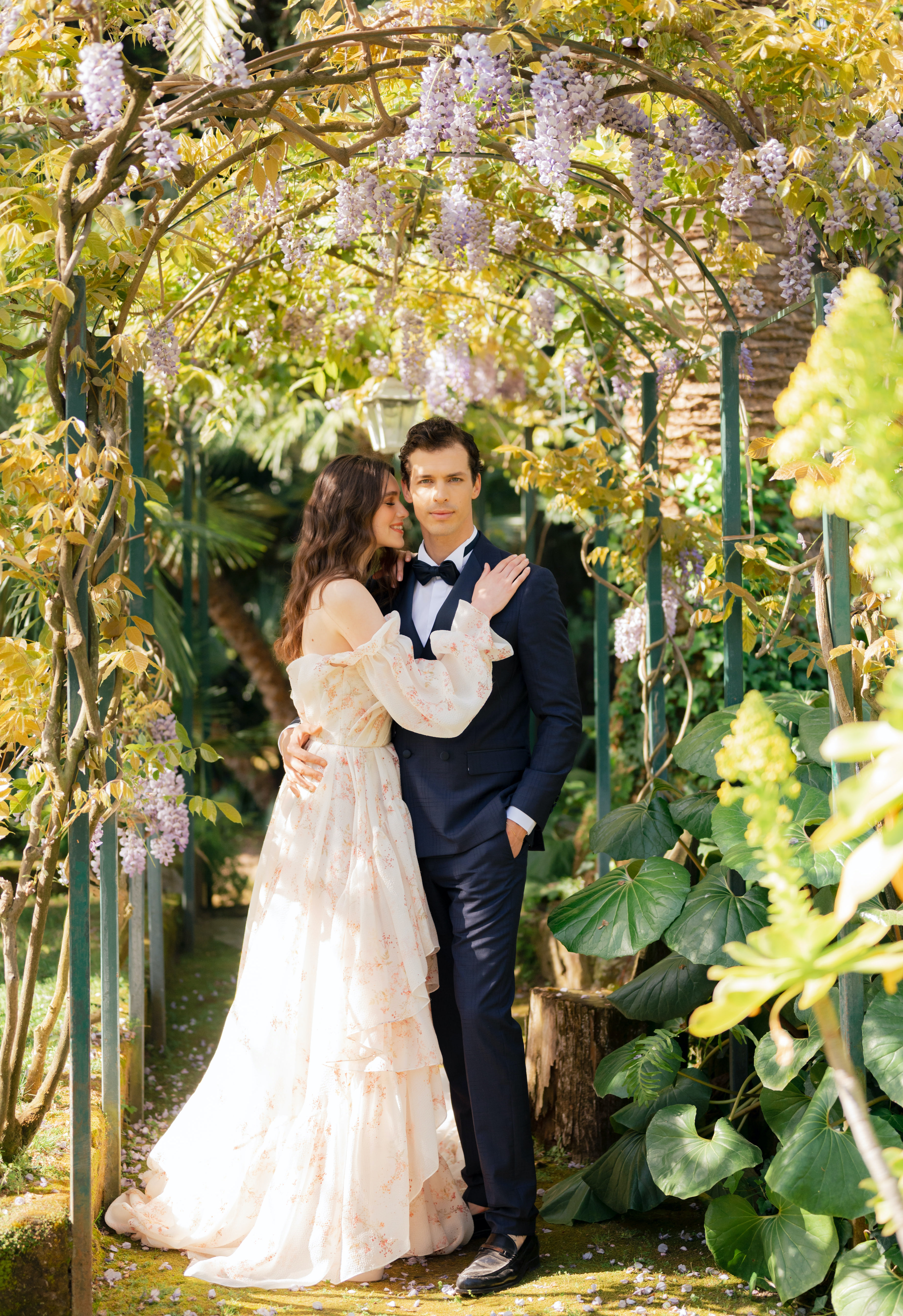 Wisteria creates a canopy of a bride and groom's head as they pose for the camera in a lush garden at their wedding venue in Italy.