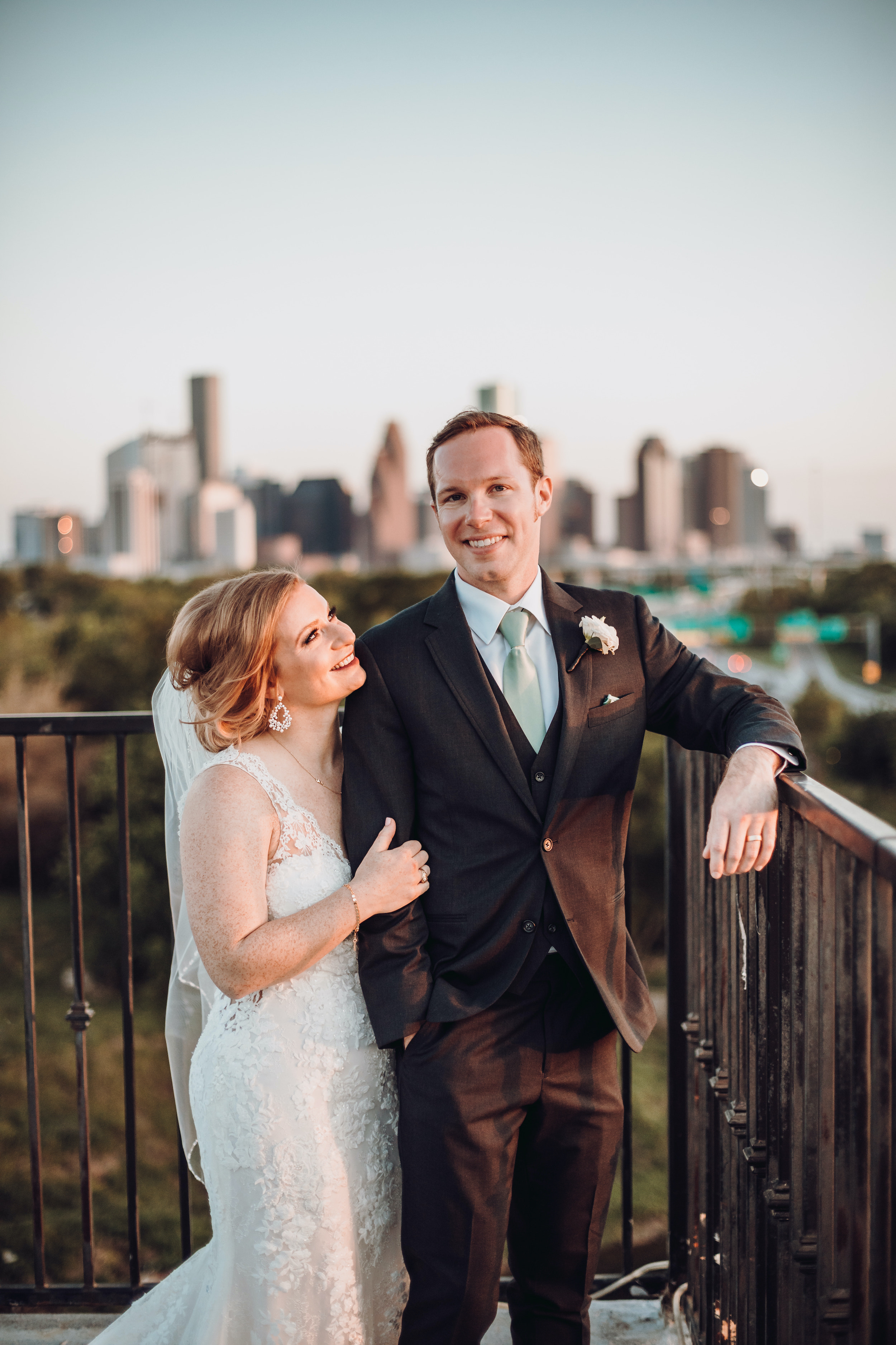 A bride and groom smile and pose on a balcony overlooking the Houston skyline.