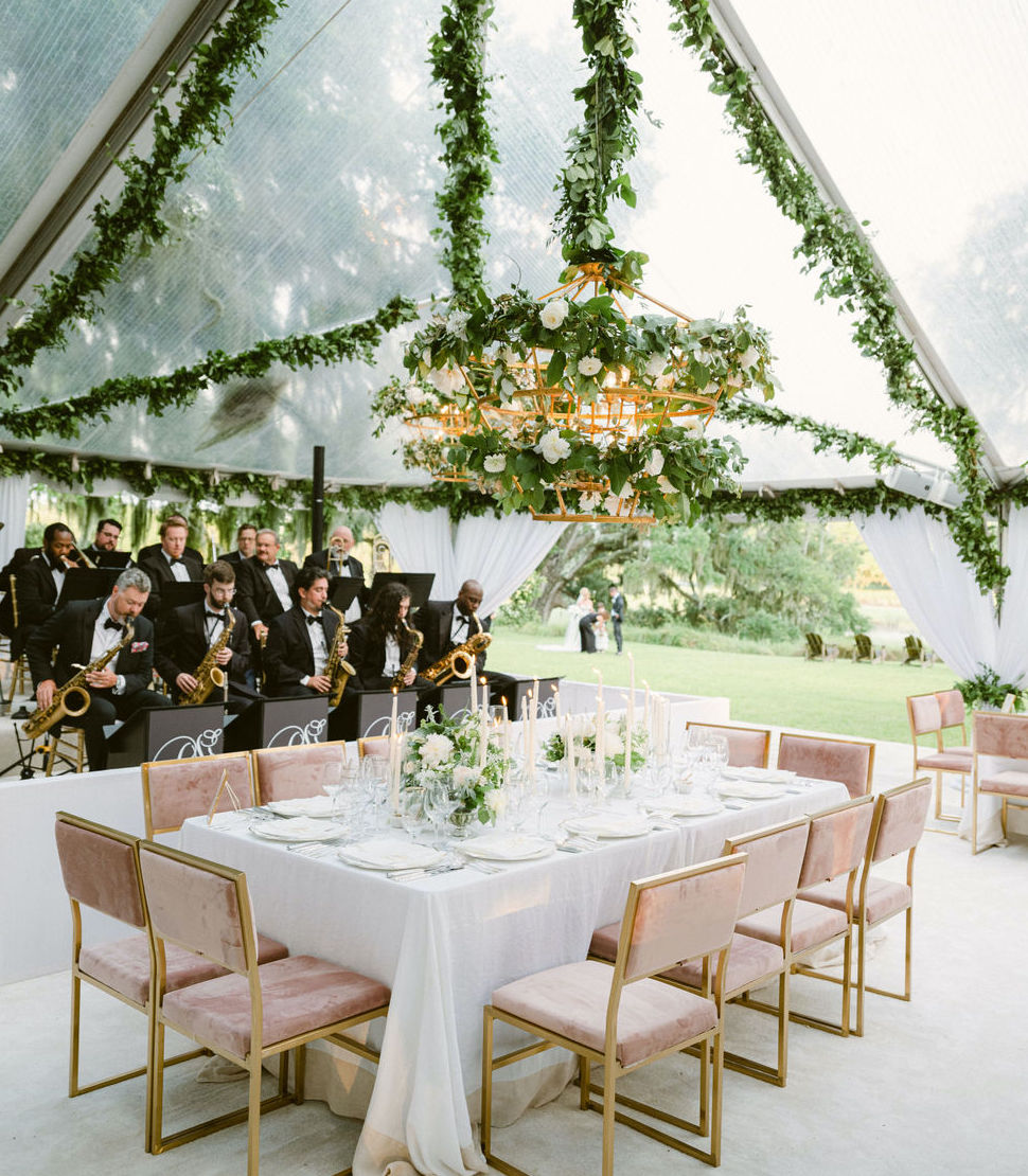 An orchestra plays under the tent for a wedding reception in the Lowcountry. The dinner tables are set with candles and white flowers and greenery in the middle of the tables.