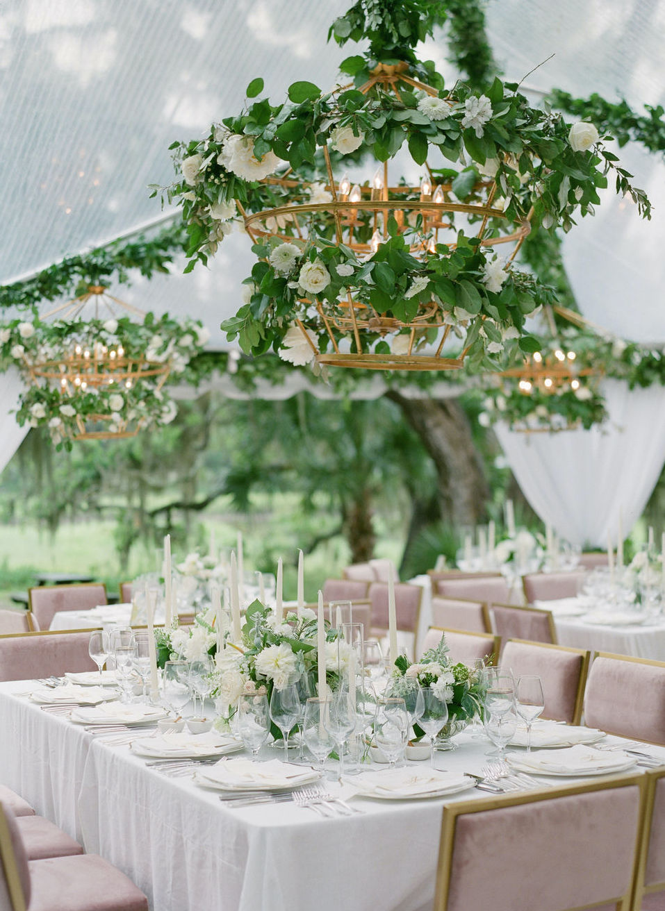 Under a wedding reception tent outside in Charleston, SC with greenery on the ceiling and in the middle of the tables. Mauve seats are set around the tables.