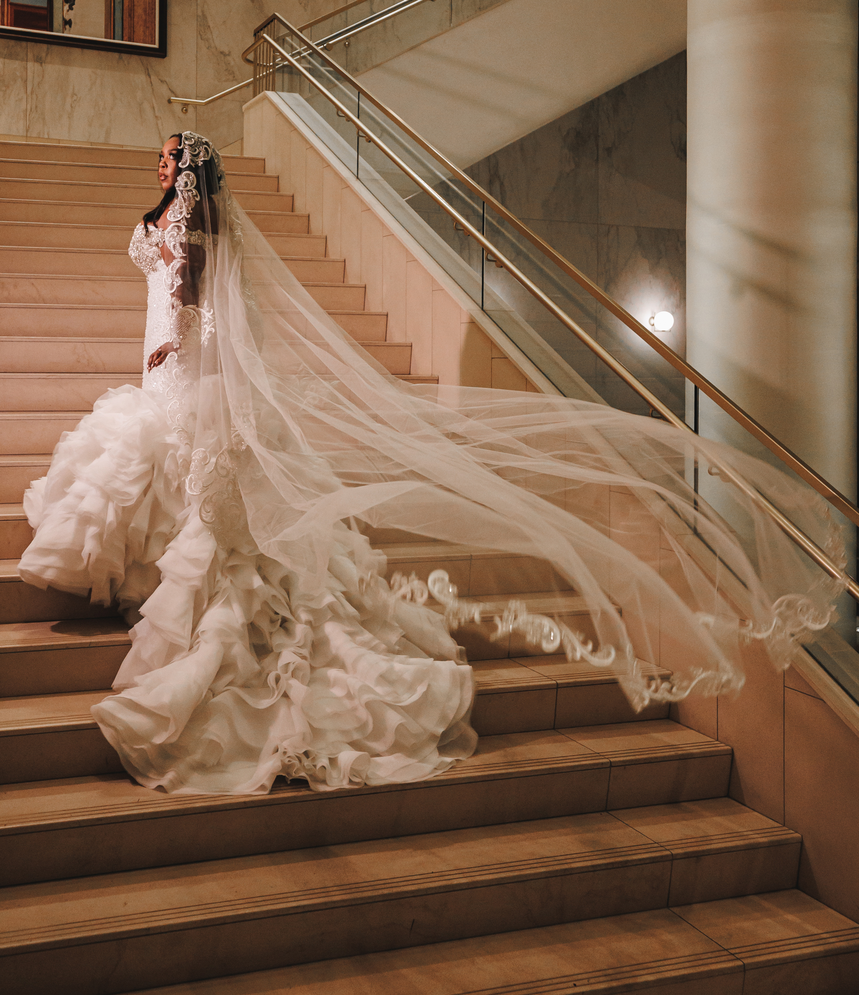 Bride, Ashley Turner, walks up a grand staircase in her wedding gown that has ruffled train and a long veil trailing behind her in the air.