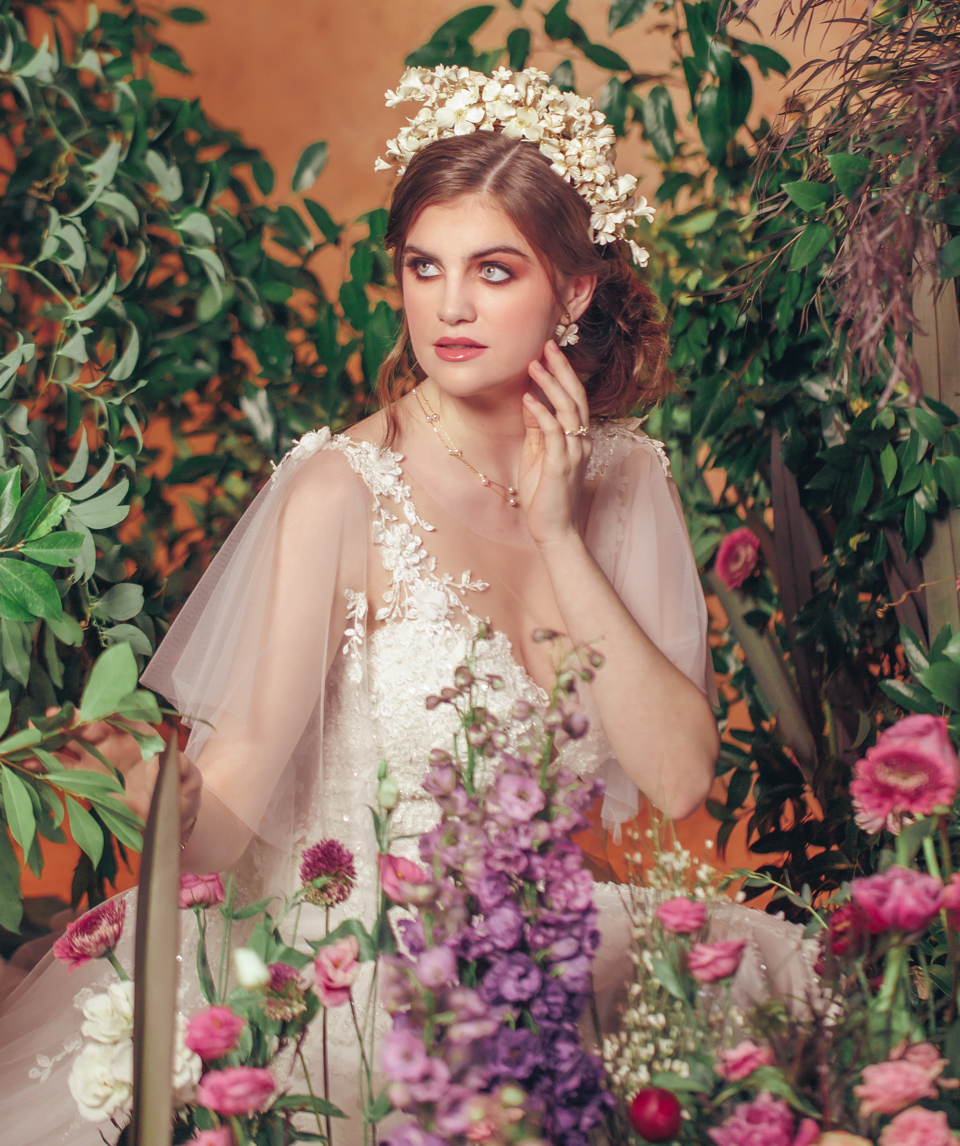 A bride wears a floral headpiece and sits in a bed of flowers.