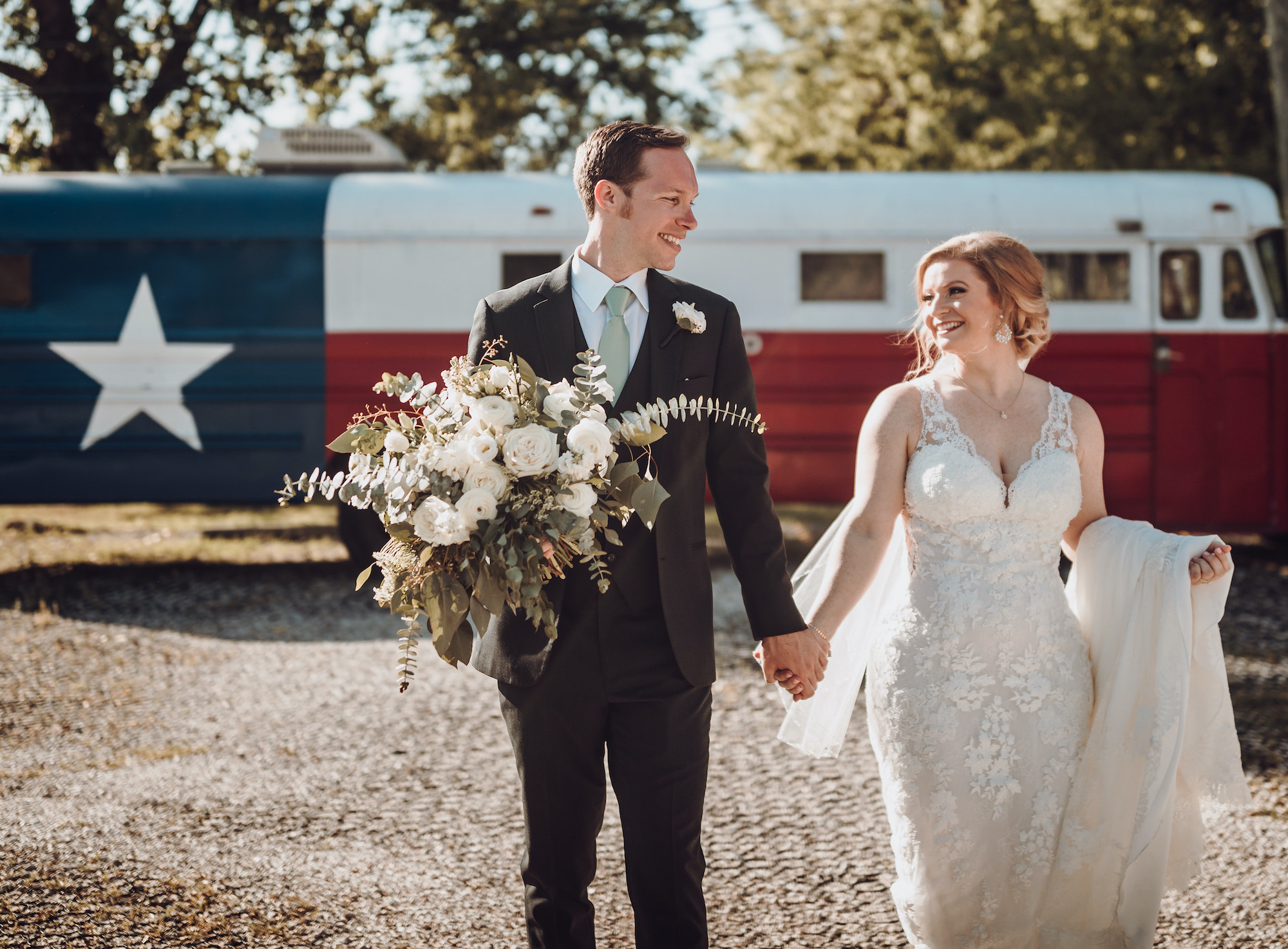 A bride and groom holds hands and walk in front of a school bus painted as a TX flag.