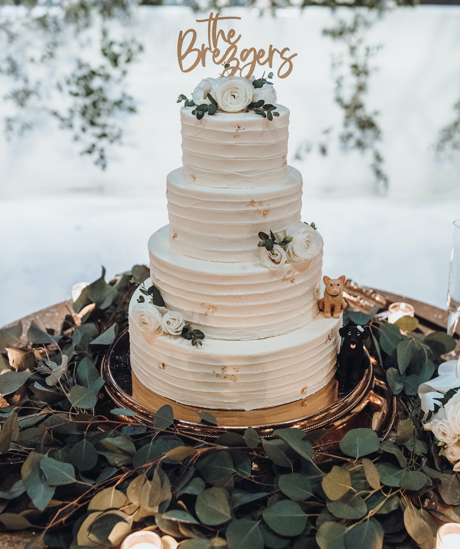 A four tiered white wedding cake sits in the center of eucalyptus leaves and has white Ranunculus flowers on it.