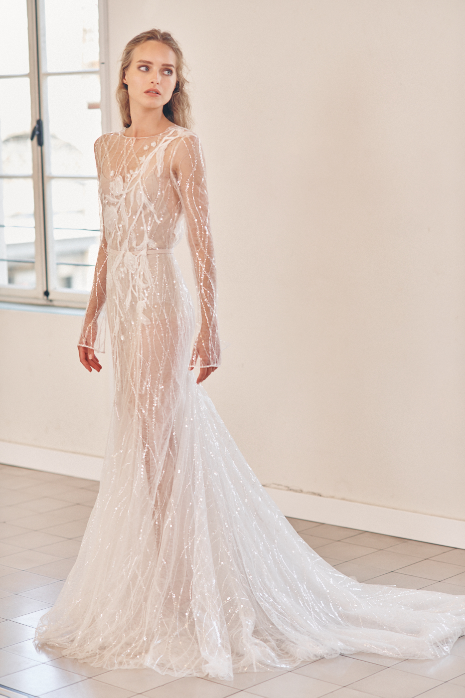 A sheer long sleeve wedding gown designed by Mira Zwillinger with shimmering flowers and a slim fit.