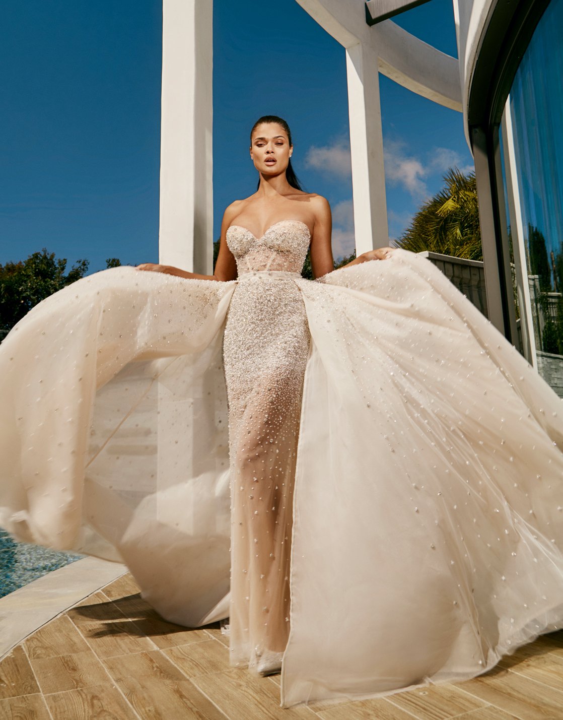 A bride holds her sparkling skirt that is blowing in the wind and she wears a Galia Lahav gown that is strapless and beige with rhinestones covering it.